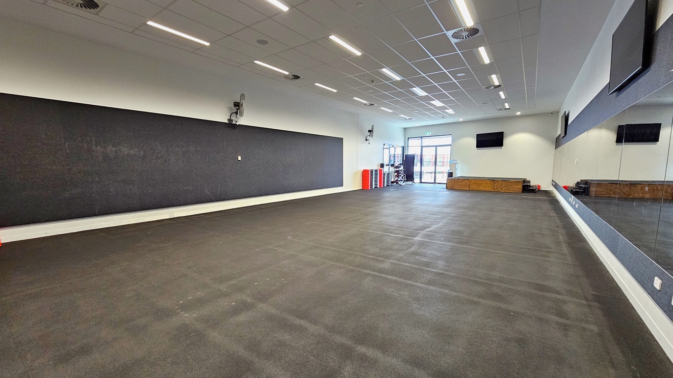 In the Fitness Room, a blackboard is on the left, and an expansive mirror is on the right. Flatscreen TVs are mounted on the upper middle and right walls, with wall fans installed on the upper left. Fitness equipment is stacked in the left corner near the glass door. The floor is covered with black rubber tiles, and the ceiling is white with fluorescent lights. In the Fitness Room, a blackboard is on the left, and an expansive mirror is on the right. Flatscreen TVs are mounted on the upper middle and right walls, with wall fans installed on the upper left. Fitness equipment is stacked in the left corner near the glass door. The floor is covered with black rubber tiles, and the ceiling is white with fluorescent lights.
