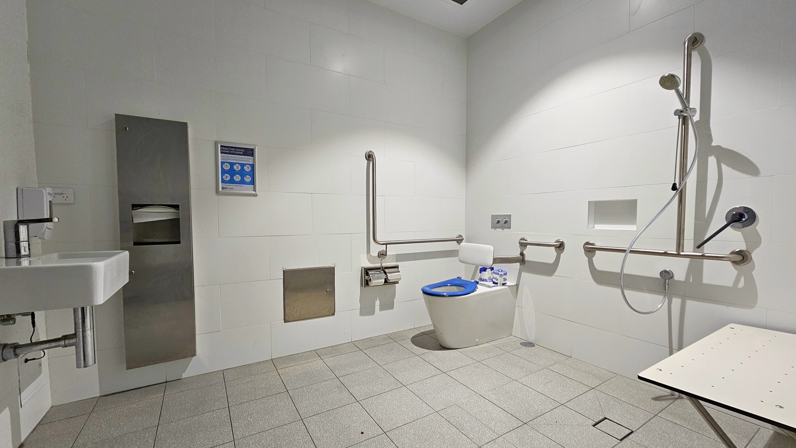 Inside the Accessible Bathroom, a toilet bowl with hand rails and a tissue dispenser is placed in the corner. To the right, a shower with handrails and a foldable bench is located. On the left, there's a washbasin next to a tissue and liquid soap dispenser. Grey tiles cover the floor, and the walls and ceiling are white. A poster is affixed to the right wall. Inside the Accessible Bathroom, a toilet bowl with hand rails and a tissue dispenser is placed in the corner. To the right, a shower with handrails and a foldable bench is located. On the left, there's a washbasin next to a tissue and liquid soap dispenser. Grey tiles cover the floor, and the walls and ceiling are white. A poster is affixed to the right wall.