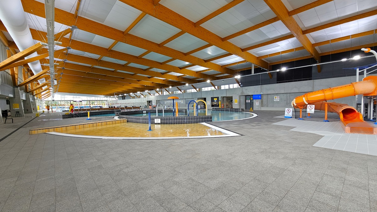 The children's splash park and leisure pool feature a beach entry, water sprays, geysers, and waterfalls. An orange slide is positioned on the right. Grey tiles surround the pool deck, and free-standing safety warnings and banners are placed on the slides. Plastic chairs are arranged on the left. The ceiling is white with wooden beams and large aircon vents. Additionally, a pool staff member is present. The children's splash park and leisure pool feature a beach entry, water sprays, geysers, and waterfalls. An orange slide is positioned on the right. Grey tiles surround the pool deck, and free-standing safety warnings and banners are placed on the slides. Plastic chairs are arranged on the left. The ceiling is white with wooden beams and large aircon vents. Additionally, a pool staff member is present.