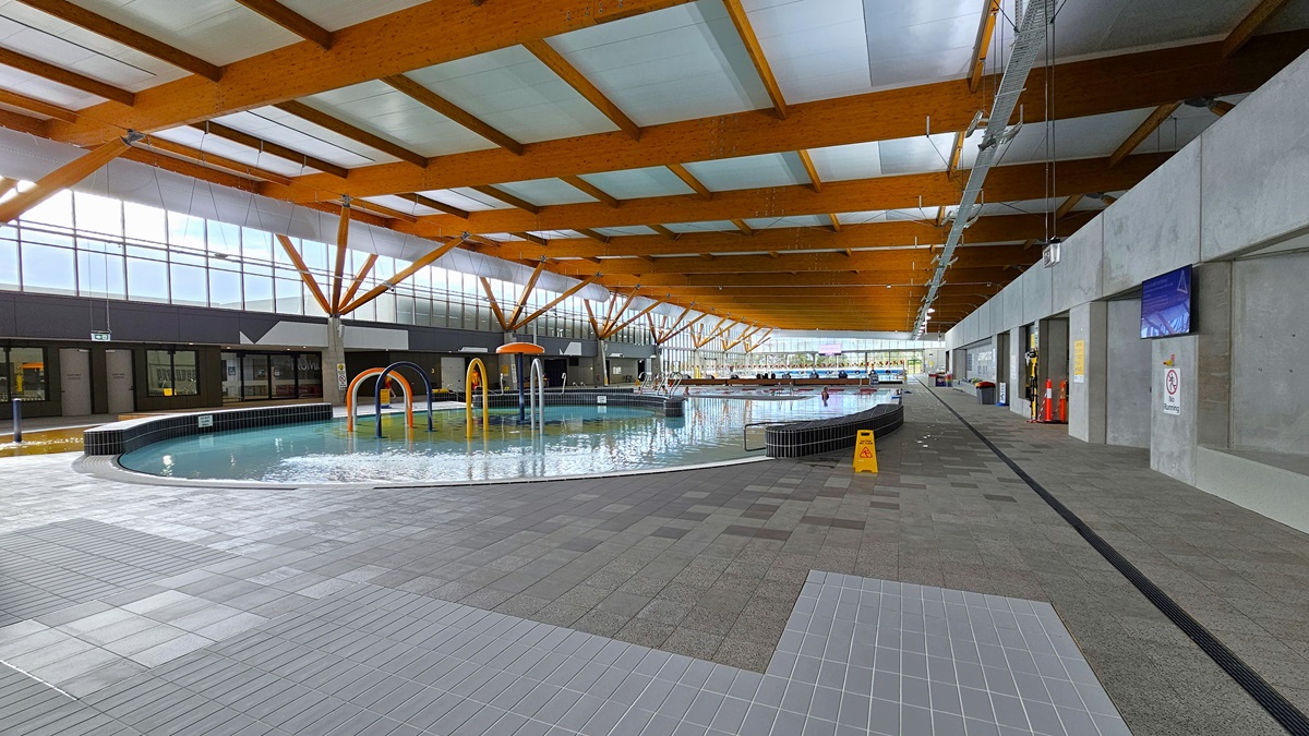 The children's splash park and leisure pool feature a beach entry, water sprays, geysers, and waterfalls. The pool has a raised deck with small black tiles, surrounded by grey checkered tiles. A yellow safety sign is visible, and a flat-screen TV is mounted on the right wall. The ceiling is embellished with wooden beams. A pool staff member is stationed beside the pool, and a glass wall surrounds the right section. The children's splash park and leisure pool feature a beach entry, water sprays, geysers, and waterfalls. The pool has a raised deck with small black tiles, surrounded by grey checkered tiles. A yellow safety sign is visible, and a flat-screen TV is mounted on the right wall. The ceiling is embellished with wooden beams. A pool staff member is stationed beside the pool, and a glass wall surrounds the right section.