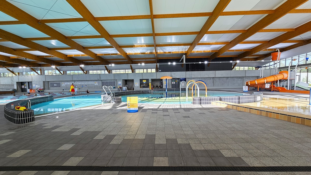 The children's splash park and leisure pool include a beach entry, water sprays, geysers, and waterfalls. The pool features a raised deck with small black tiles, bordered by grey checkered tiles. A yellow safety sign is visible. An orange slide with a tipping bucket is situated on the right, near the glass wall. The ceiling is adorned with wooden beams, and a pool staff member is stationed beside the pool. The children's splash park and leisure pool include a beach entry, water sprays, geysers, and waterfalls. The pool features a raised deck with small black tiles, bordered by grey checkered tiles. A yellow safety sign is visible. An orange slide with a tipping bucket is situated on the right, near the glass wall. The ceiling is adorned with wooden beams, and a pool staff member is stationed beside the pool.