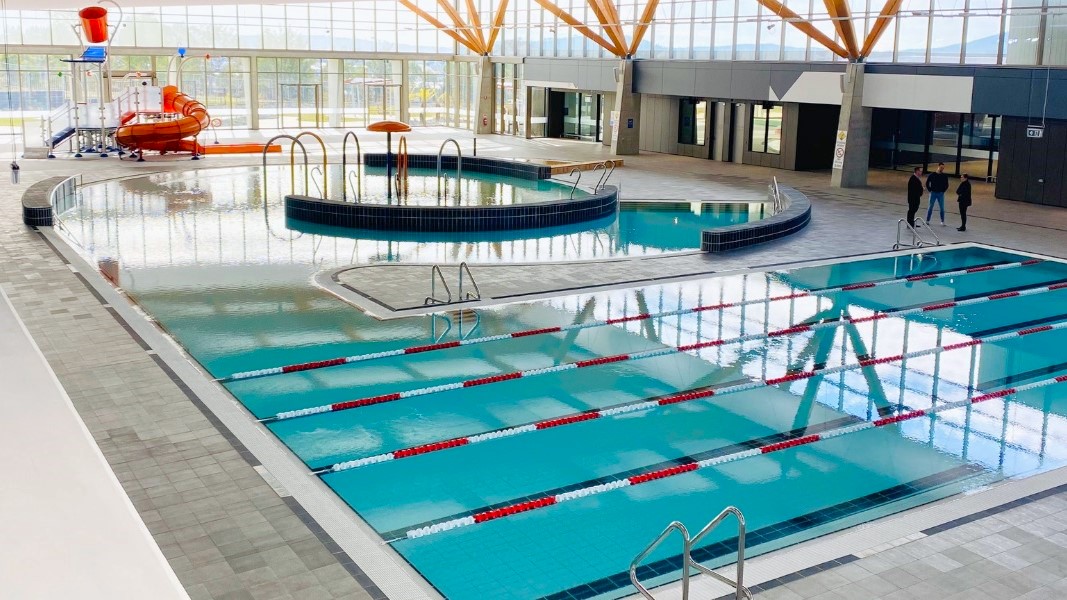 The training pool features handrails and floating ropes. Connected to it is the children's splash park and leisure pool, equipped with a beach entry, water sprays, geysers, and waterfalls. The pool has a raised deck with small black tiles, bordered by grey checkered tiles. Furthermore, a slide with a tipping bucket is positioned near the glass wall. People are standing beside the pool on the right. The training pool features handrails and floating ropes. Connected to it is the children's splash park and leisure pool, equipped with a beach entry, water sprays, geysers, and waterfalls. The pool has a raised deck with small black tiles, bordered by grey checkered tiles. Furthermore, a slide with a tipping bucket is positioned near the glass wall. People are standing beside the pool on the right.