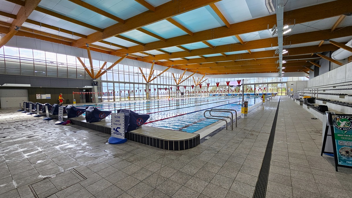 The 50m Heated Swimming Pool has handrails, floating ropes, and banners. Diving boards, wrapped with blue covers, are aligned on the raised pool deck on the left. Free-standing safety banners are placed on the pool deck. On the right, there is tiered seating with additional free-standing banners. The ceiling is adorned with wooden beams and floodlights. The floor is covered with grey tiles, featuring an overflow grid. A glass wall surrounds the pool. The 50m Heated Swimming Pool has handrails, floating ropes, and banners. Diving boards, wrapped with blue covers, are aligned on the raised pool deck on the left. Free-standing safety banners are placed on the pool deck. On the right, there is tiered seating with additional free-standing banners. The ceiling is adorned with wooden beams and floodlights. The floor is covered with grey tiles, featuring an overflow grid. A glass wall surrounds the pool.
