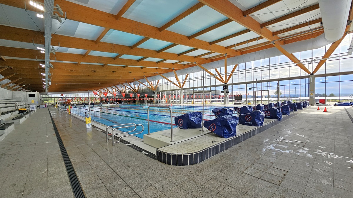 The 50m Heated Swimming Pool features handrails, floating ropes, and orange banners. Diving boards, covered in blue, are aligned on the raised pool deck on the right. Safety cones and banners are visible on the pool deck. The ceiling is adorned with wooden beams and floodlights. The floor is covered with grey tiles, including an overflow grid. The pool is surrounded by a glass wall. The 50m Heated Swimming Pool features handrails, floating ropes, and orange banners. Diving boards, covered in blue, are aligned on the raised pool deck on the right. Safety cones and banners are visible on the pool deck. The ceiling is adorned with wooden beams and floodlights. The floor is covered with grey tiles, including an overflow grid. The pool is surrounded by a glass wall.