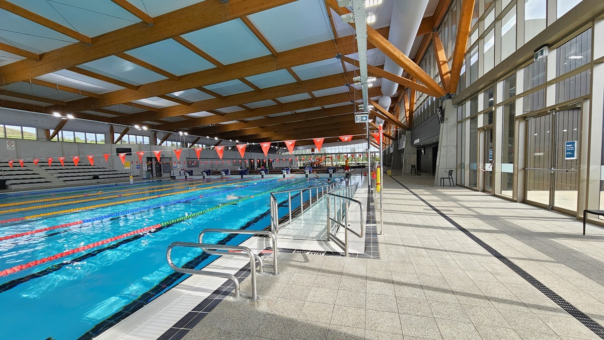 The 50m Heated Swimming Pool includes a ramp with handrails, floating ropes, and orange banners. At the pool's end, diving boards, covered in blue, align with the raised pool deck. To the right, a glass wall and door are present. The ceiling features wooden beams, large aircon vents, and floodlights. The floor is tiled in grey, incorporating an overflow grid. To the right is a glass wall and a glass door. The 50m Heated Swimming Pool includes a ramp with handrails, floating ropes, and orange banners. At the pool's end, diving boards, covered in blue, align with the raised pool deck. To the right, a glass wall and door are present. The ceiling features wooden beams, large aircon vents, and floodlights. The floor is tiled in grey, incorporating an overflow grid. To the right is a glass wall and a glass door.