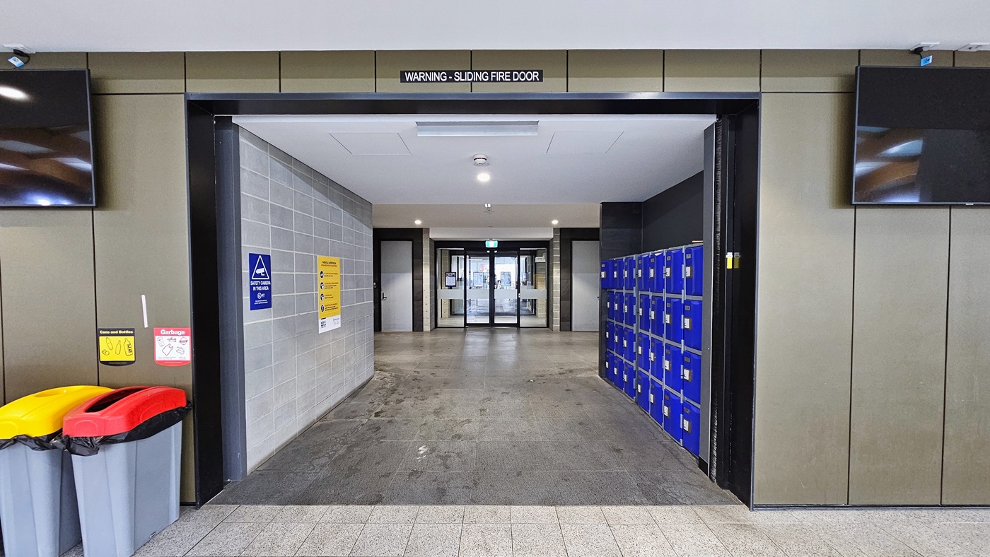 Blue lockers are situated on the right, while the grey brick wall opposite displays posters and safety signs. Outside, two rubbish bins with segregation signs are placed on the left, beneath a flatscreen TV. Another flatscreen TV is mounted on the right wall. The floor transitions from light to dark grey, and the ceiling is uniformly white. Further in, there is a glass door entry. Blue lockers are situated on the right, while the grey brick wall opposite displays posters and safety signs. Outside, two rubbish bins with segregation signs are placed on the left, beneath a flatscreen TV. Another flatscreen TV is mounted on the right wall. The floor transitions from light to dark grey, and the ceiling is uniformly white. Further in, there is a glass door entry.