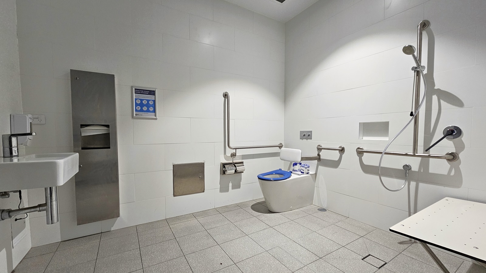 Within the accessible bathroom, a toilet bowl equipped with hand grabs and a tissue dispenser is positioned in the corner. To its right, there is a shower with handrails, accompanied by a foldable bench. On the left is a washbasin, adjacent to a tissue and liquid soap dispenser. The floor is covered with grey tiles, and the walls and ceiling are white. Within the accessible bathroom, a toilet bowl equipped with hand grabs and a tissue dispenser is positioned in the corner. To its right, there is a shower with handrails, accompanied by a foldable bench. On the left is a washbasin, adjacent to a tissue and liquid soap dispenser. The floor is covered with grey tiles, and the walls and ceiling are white.