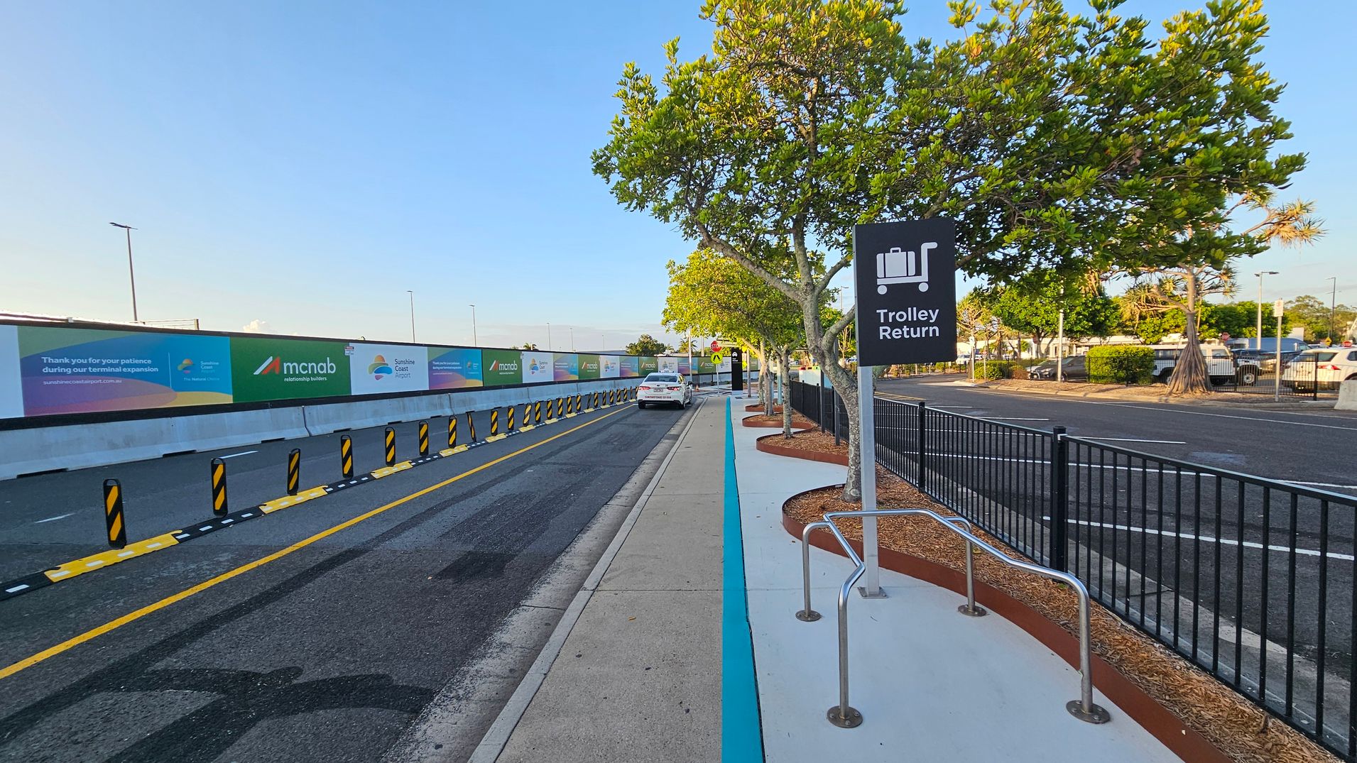 A road with a designated lane on the left, marked by yellow lines and black-and-yellow bollards. A white car is parked further down the lane. On the right, a pavement with a "Trolley Return" sign and metal trolley racks. Trees with green foliage line the pavement, and a black metal fence separates the pavement from a car park. Advertising banners are displayed on a wall to the left. The ground is asphalt and concrete, with mulch around the tree bases.