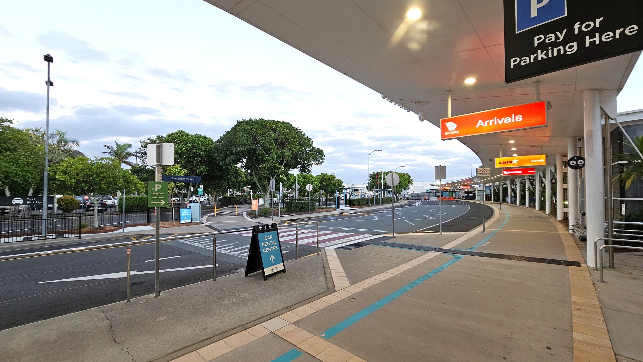 A covered walkway at an airport with a beige tiled floor and a white ceiling with lights. On the right, there are signs for "Arrivals" in orange and "Pay for Parking Here" in blue. The road curves to the left with marked lanes and pedestrian crossings. On the left, there are trees and signs for "Passenger Pick-up" and "Car Rental Centre". A fence separates the walkway from the road.