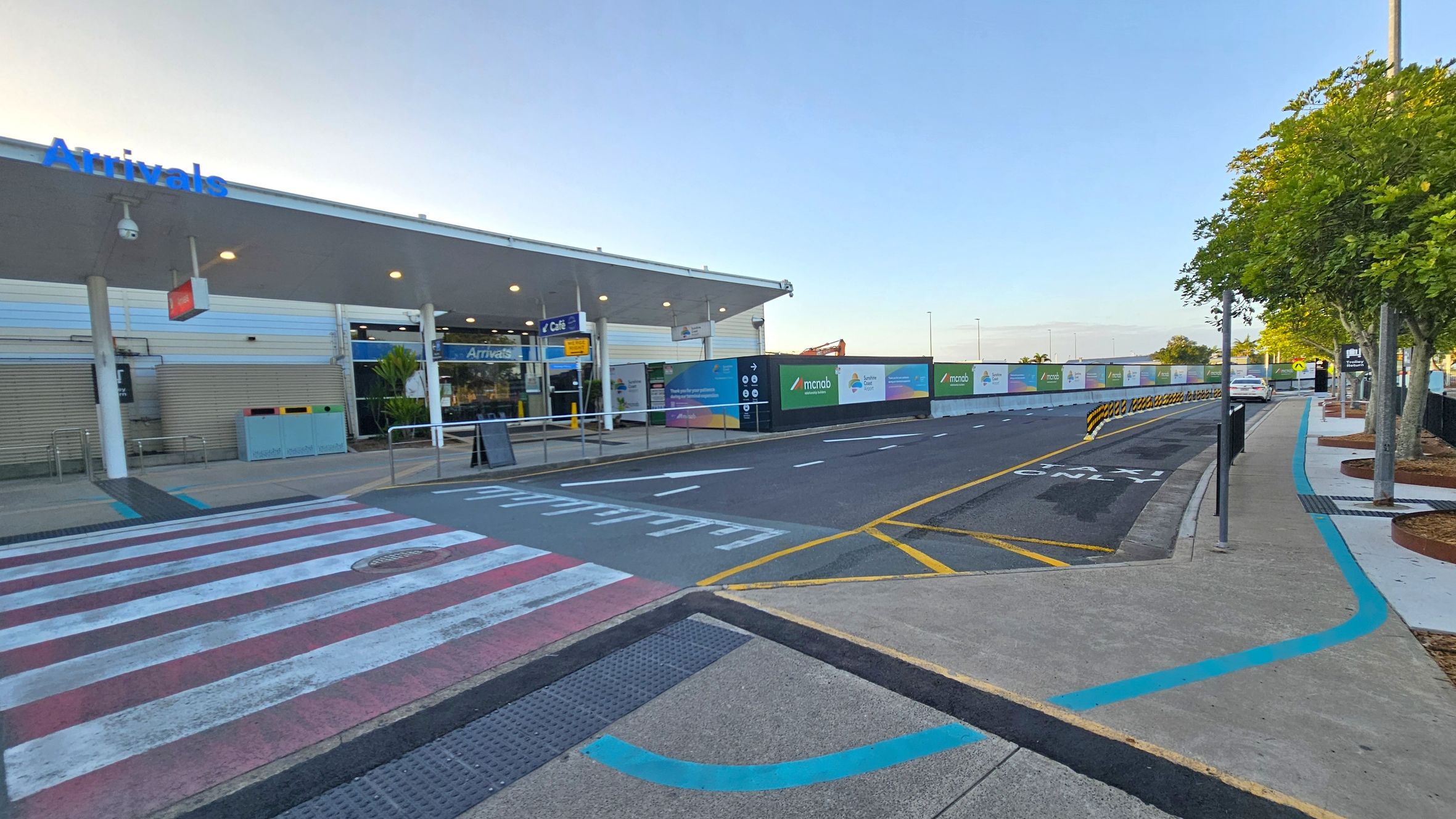 On the left, a building with a sign reading "Arrivals" and a café sign is visible. The building has a covered entrance with glass doors. In the middle, a road with marked lanes and a pedestrian crossing is present. On the right, there is a footpath lined with trees and a parked car. Construction barriers with advertisements are along the road. The ground is paved with concrete and asphalt, and the ceiling of the building's entrance is white with lights.