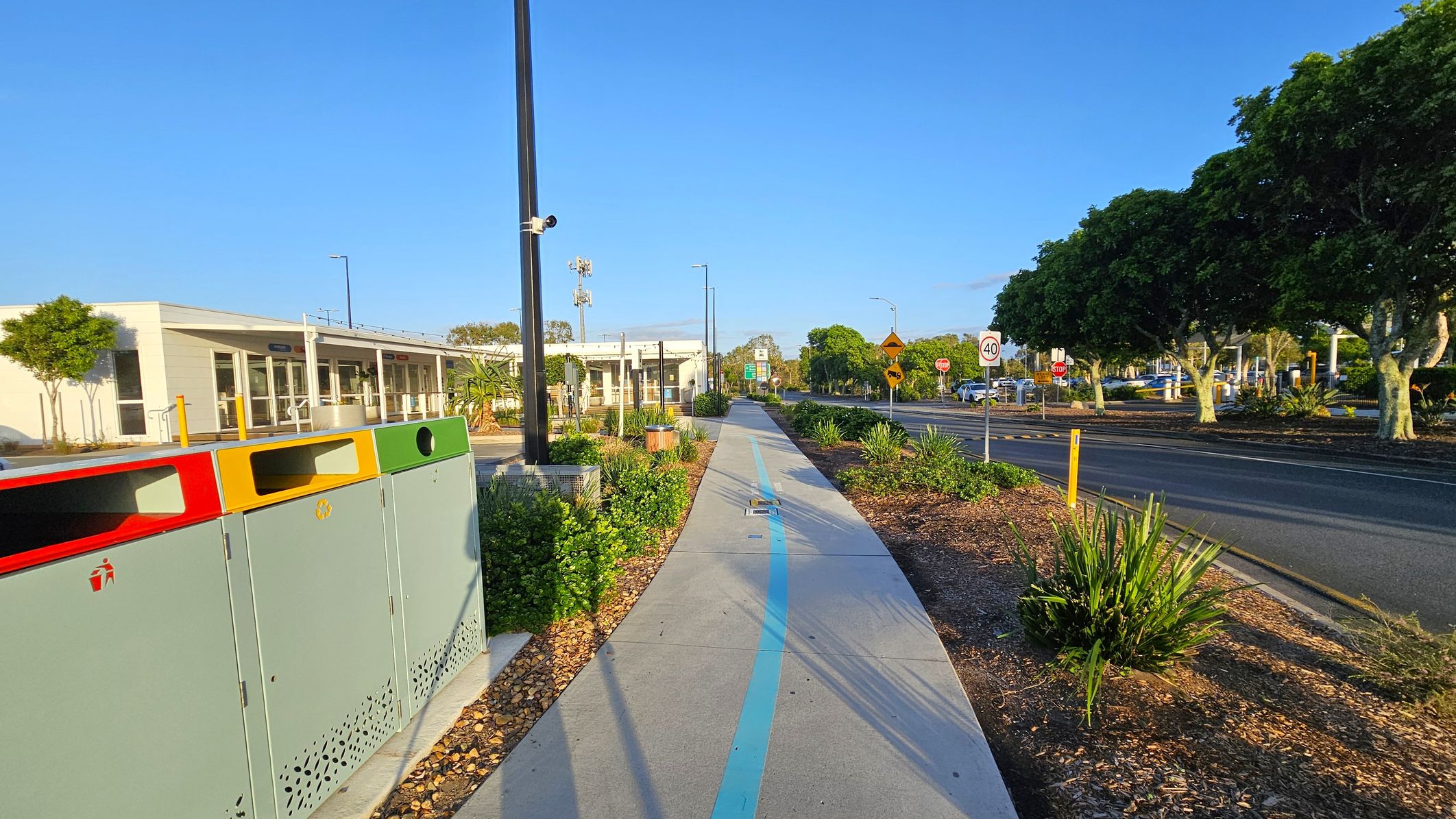 A pathway runs through the centre, marked with a blue line. On the left, there are colourful bins and a modern building with large windows. On the right, a road with a 40 km/h speed limit sign is lined with lush green trees. The foreground features landscaped plants and a tall streetlight. The background shows more trees and a clear sky.