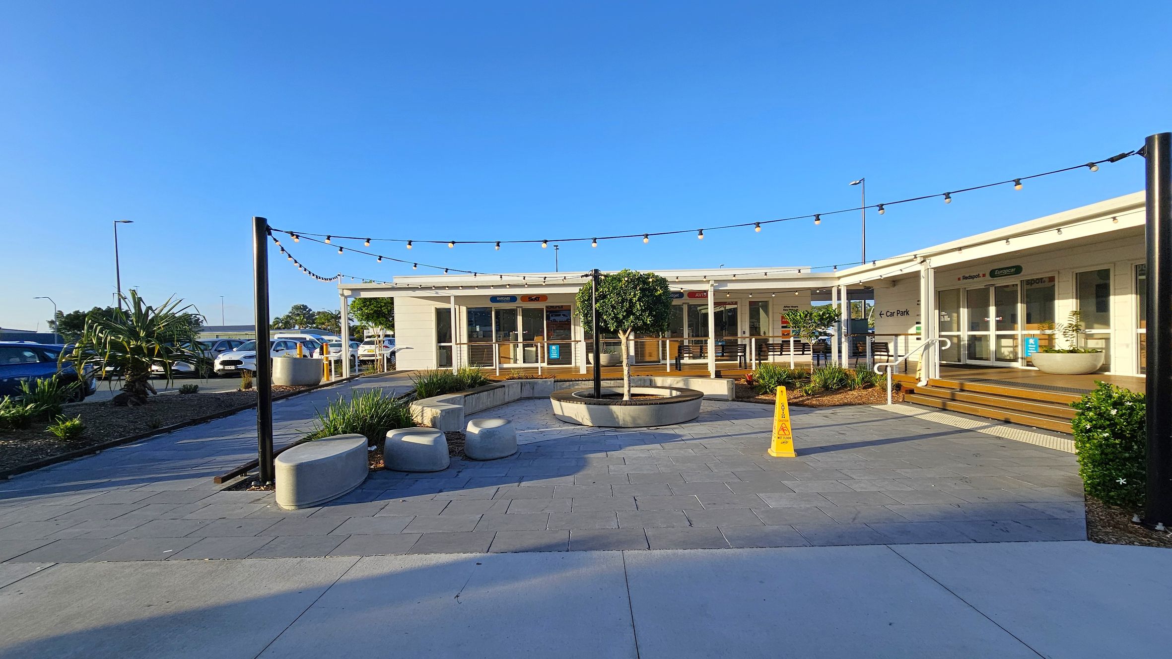 A courtyard with a paved area featuring a circular planter with a small tree in the centre. Surrounding the planter are concrete benches. Overhead, string lights are suspended between black poles. To the right, a building with glass doors and steps leads to a car park. A yellow caution sign is visible near the steps. On the left, there are parked cars and some greenery, including a large plant. The sky is clear and blue.