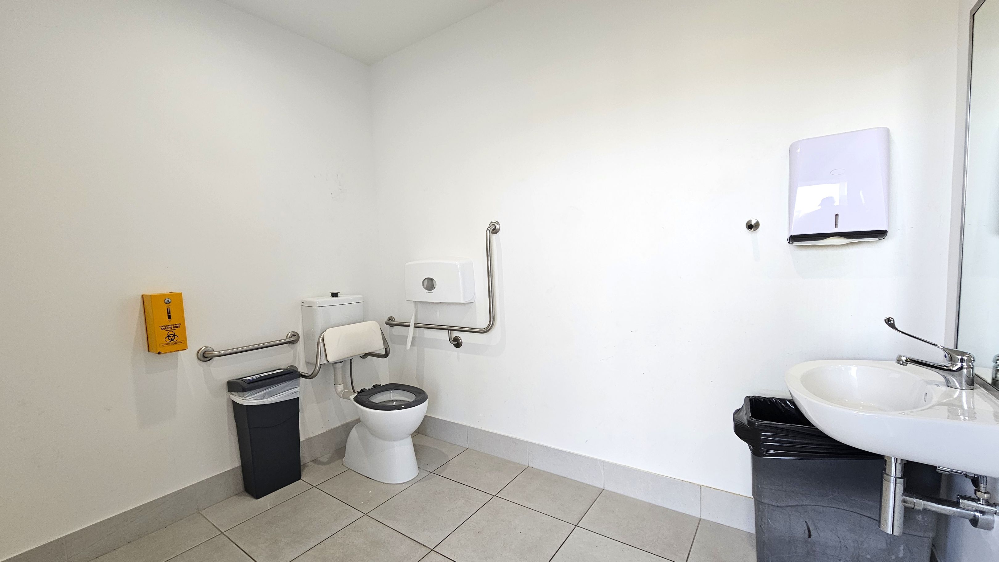 A clean, accessible bathroom with a white toilet featuring support rails on both sides. To the left, a yellow emergency assistance box is mounted on the wall above a black bin. The floor is tiled in light grey. On the right, a white sink with a lever tap is positioned above another black bin. A paper towel dispenser is mounted on the wall above the sink. The walls are plain white, and the space is well-lit.