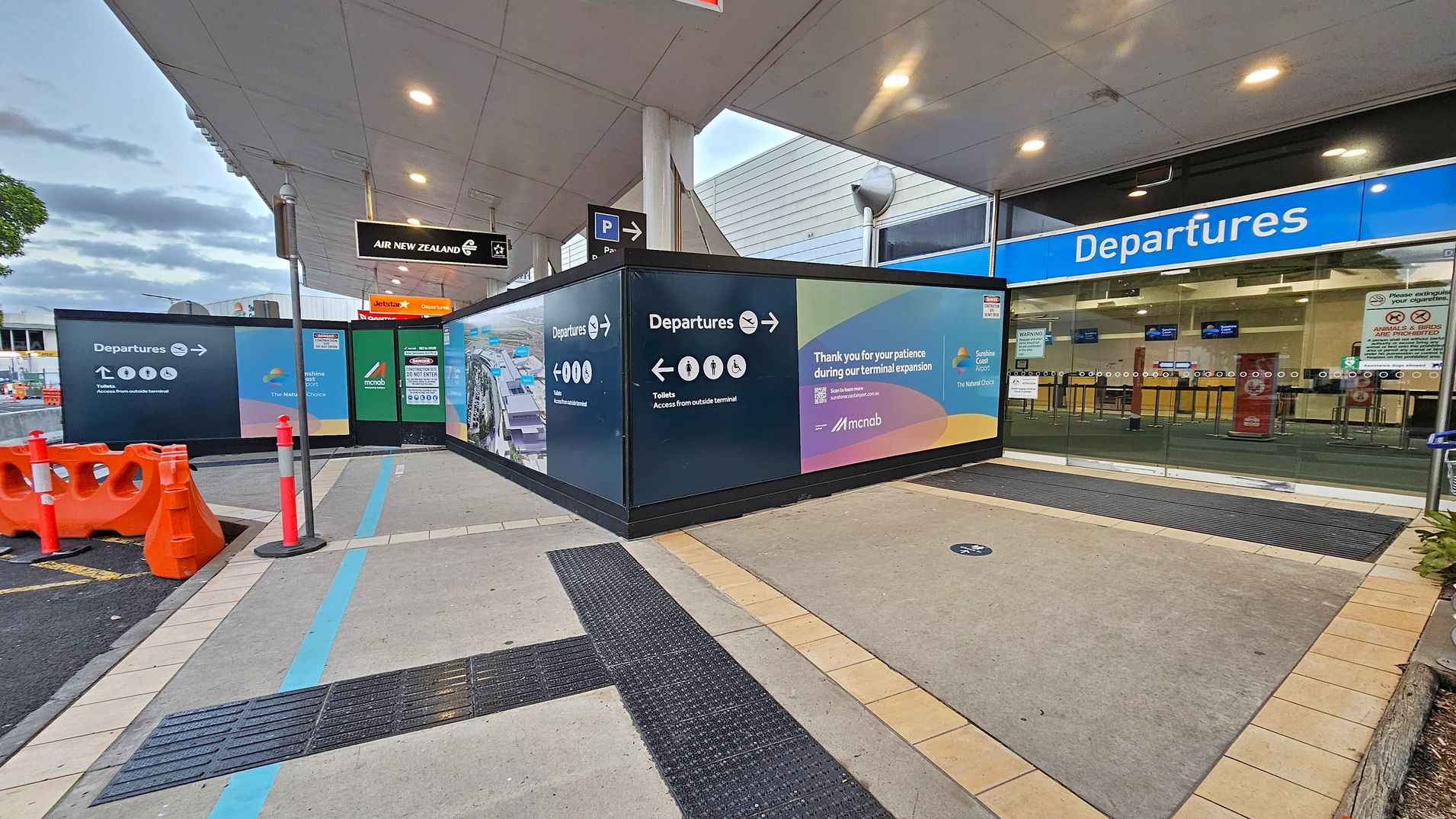 On the left, there are orange traffic barriers and a sign for Air New Zealand. In the centre, a construction wall displays directions to departures and toilets, along with a message thanking for patience during terminal expansion. On the right, the entrance to the departures area is visible with a blue sign above glass doors. The floor is a mix of concrete and tiled sections, and the ceiling is white with recessed lighting.