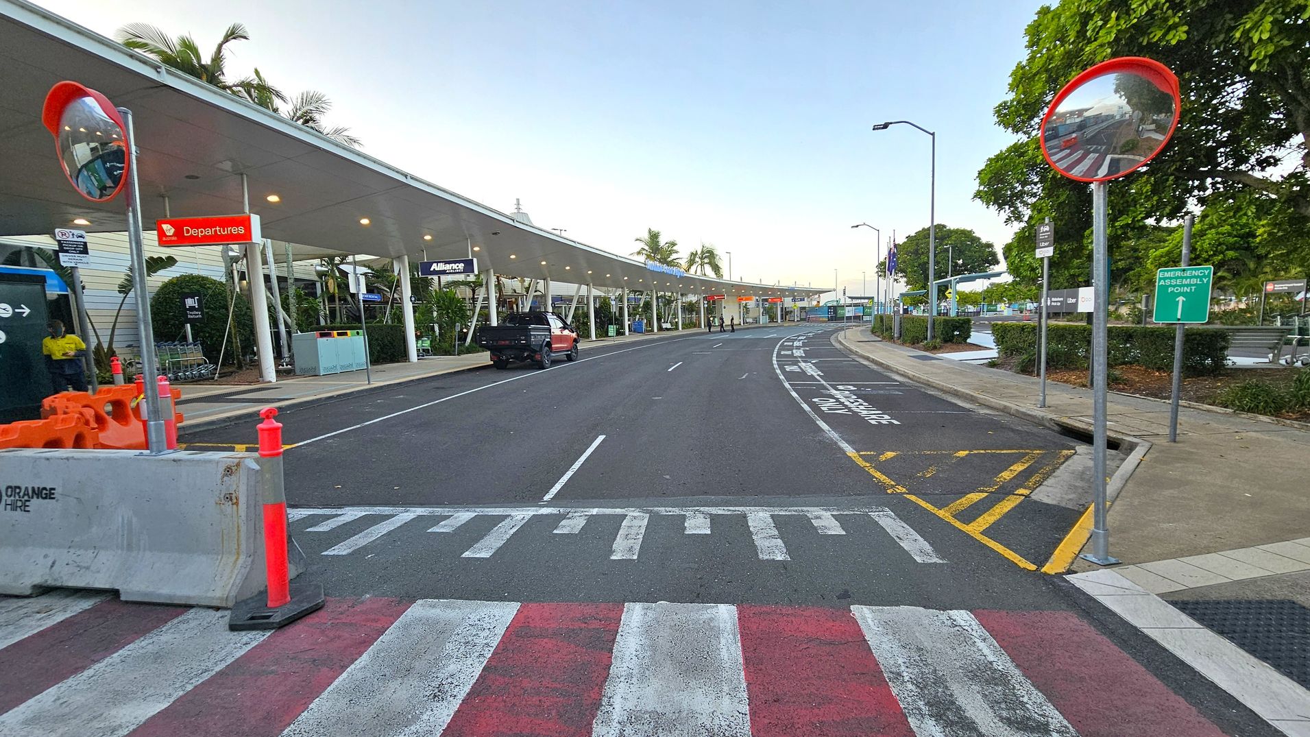 A road with a pedestrian crossing in the foreground, marked with red and white stripes. On the left, there are orange traffic cones and a concrete barrier. A covered walkway with signs for "Departures" and "Alliance" runs along the left side, with a black vehicle parked nearby. On the right, a convex mirror and an "Emergency Assembly Point" sign are visible. Trees and bushes line the pavement, and the road curves into the distance.