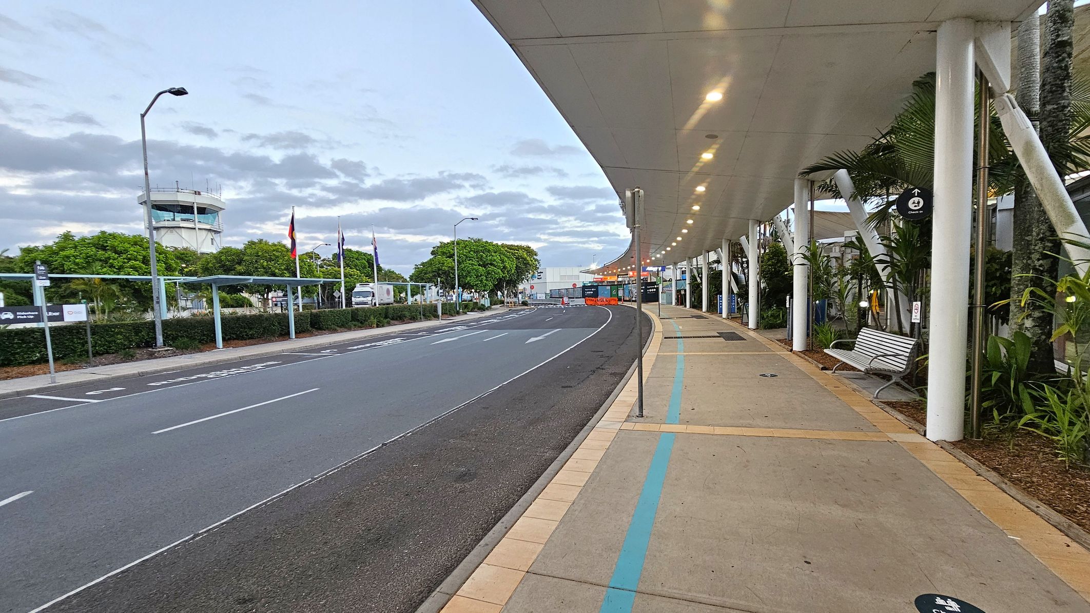 A curved road runs through the centre, with a pedestrian walkway on the right. The walkway is covered by a white ceiling with lights and supported by white pillars. A bench and some plants are along the walkway. On the left, there are trees and several flags near a fence. A control tower is visible in the background. The road has marked lanes and a sign indicating a pick-up area. The scene is well-lit, suggesting early morning or late afternoon.
