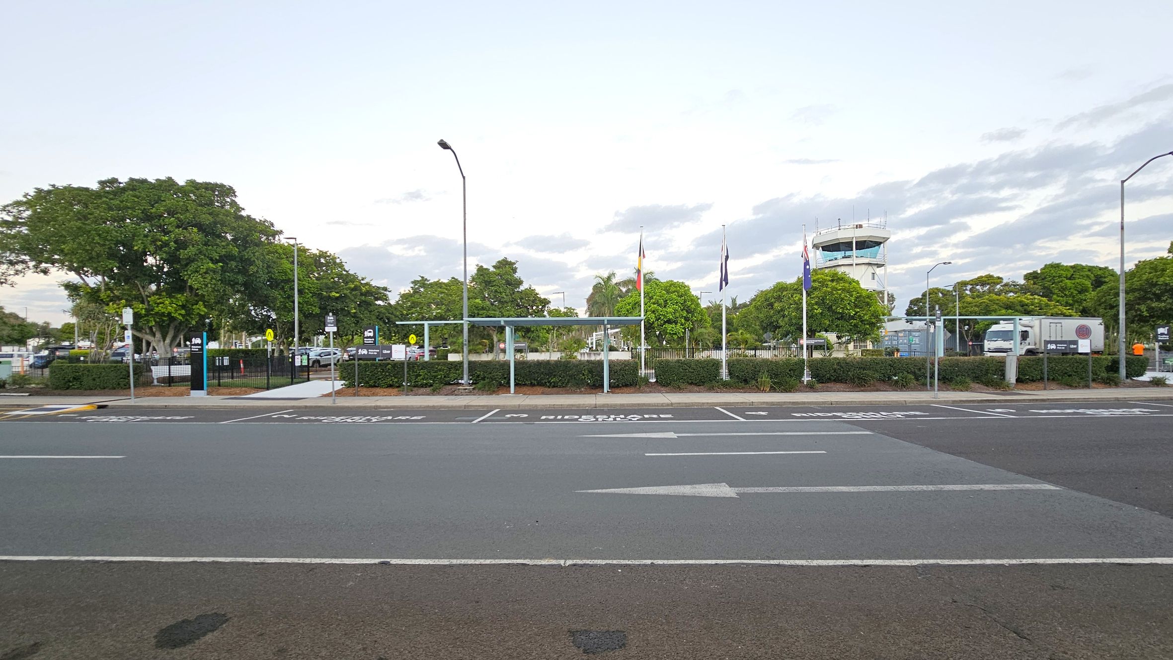 A road with marked parking spaces in the foreground, labelled for rideshare and passenger pick-up. Behind the road, there is a fenced area with a bus shelter and several flagpoles displaying flags. Trees and shrubs are scattered throughout the area. In the background, a control tower is visible, along with a parked truck. Street lamps are positioned along the road and within the fenced area.