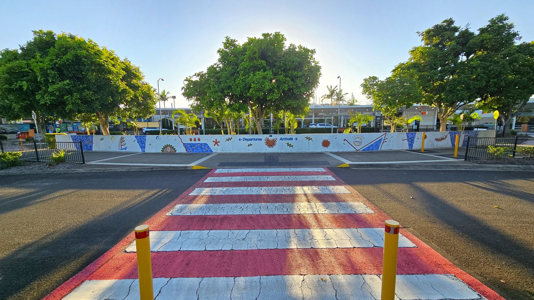 A red and white striped pedestrian crossing leads to a painted wall with colourful designs and arrows indicating "Departures" and "Arrivals". Behind the wall, there are several large green trees and a few smaller palm trees. The ground is asphalt, and there are yellow bollards near the crossing. The area is bordered by black metal fencing on both sides. In the background, there are buildings partially visible through the trees.