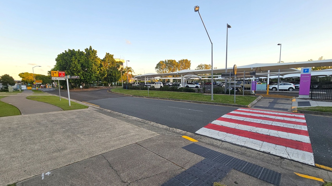A road with a red and white striped pedestrian crossing in the foreground. To the left, there's a footpath with grass and a sign that reads "PEAKER STOP DO NOT OVERTAKE." In the background, a parking area with several cars is visible, covered by a white canopy. Trees and streetlights are scattered throughout the scene, and a purple sign indicating "Covered" parking is on the right. The sky is clear, and the area is well-lit.