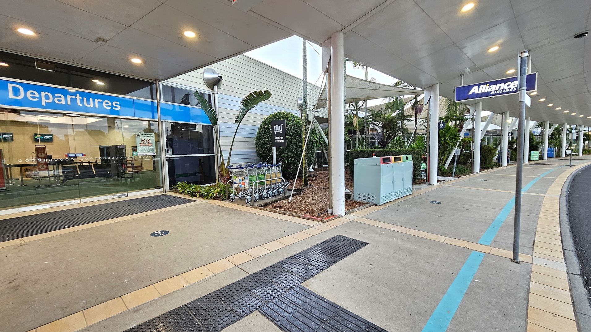 Entrance to an airport terminal with a blue "Departures" sign above glass sliding doors on the left. A row of luggage trolleys is parked near a "Trolley Return" sign. The right side features a covered walkway with a sign for "Alliance Airlines" and a row of rubbish bins. The ground is paved with a mix of concrete and tiles, and there are plants and small trees along the building's exterior. The ceiling is lined with lights.