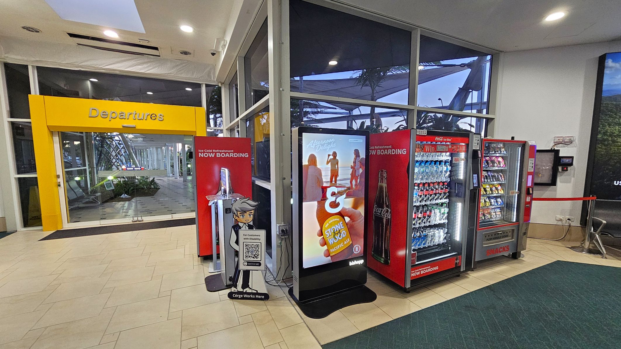 A brightly lit airport area with a yellow "Departures" sign above glass doors on the left. In the centre, there's a digital display with an advertisement, flanked by two red vending machines stocked with drinks and snacks. A stand with a cartoon character and a QR code is in front of the display. The floor is tiled in a light colour, and the ceiling has recessed lighting. Large windows in the background reveal some outdoor structures and plants.
