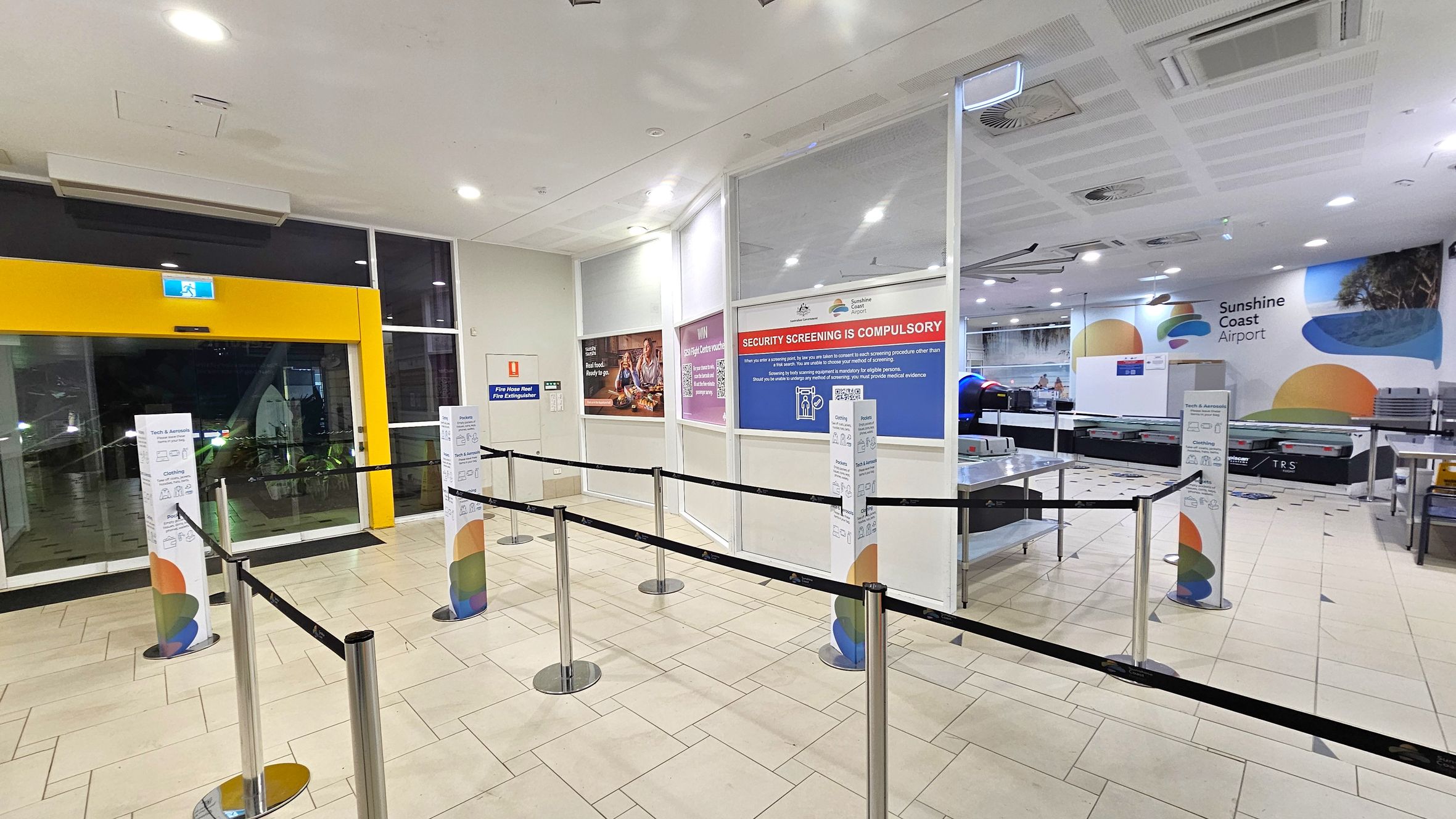 A spacious airport security area with a tiled floor. On the left, a yellow-framed glass exit door with an illuminated exit sign above. In the centre, retractable barriers guide passengers, with informational signs on pillars. On the right, a security screening area with a conveyor belt and a large wall sign reading "Sunshine Coast Airport." The ceiling has recessed lighting and air vents. Walls display various informational posters.