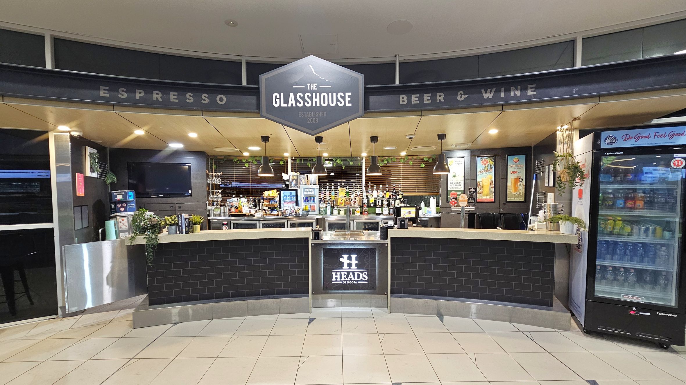 A bar area with a sign reading "The Glasshouse" above. The counter is curved with a black tile front and a "Heads" logo. Behind the counter, there are various bottles and a coffee machine. To the right, a glass-fronted refrigerator displays drinks. The ceiling has recessed lighting and hanging lamps. The floor is tiled in a light colour. Potted plants are placed on the counter and hanging on the right.