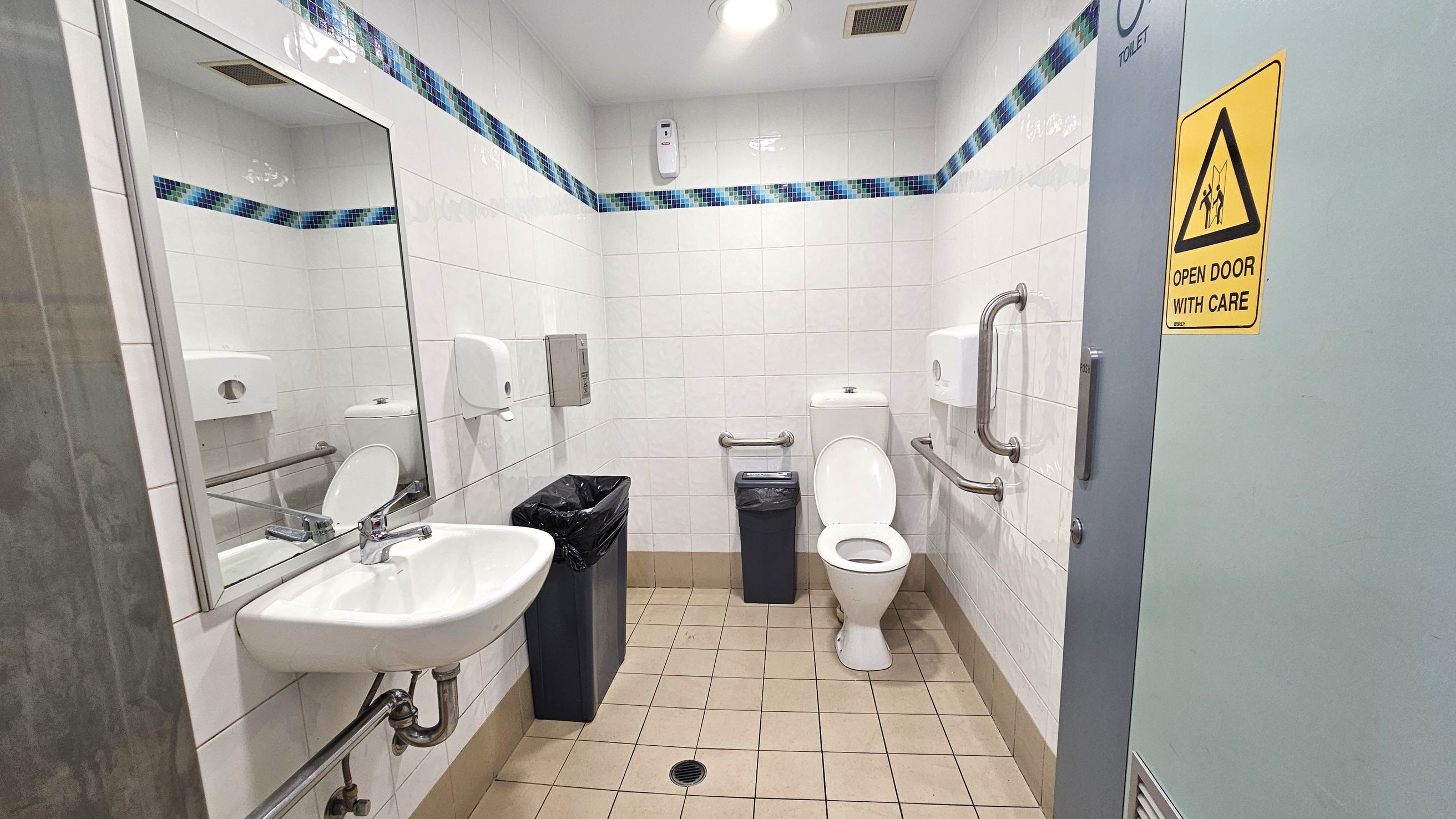 A public restroom with white tiled walls and a blue mosaic border. On the left, a wall-mounted sink with a mirror above it. Below the sink, there are exposed pipes and a metal support bar. In the centre, a white toilet with grab bars on either side and a small bin next to it. On the right, a door with a yellow caution sign reading "Open Door With Care." The floor is covered with beige tiles, and the ceiling has a light fixture and a vent.
