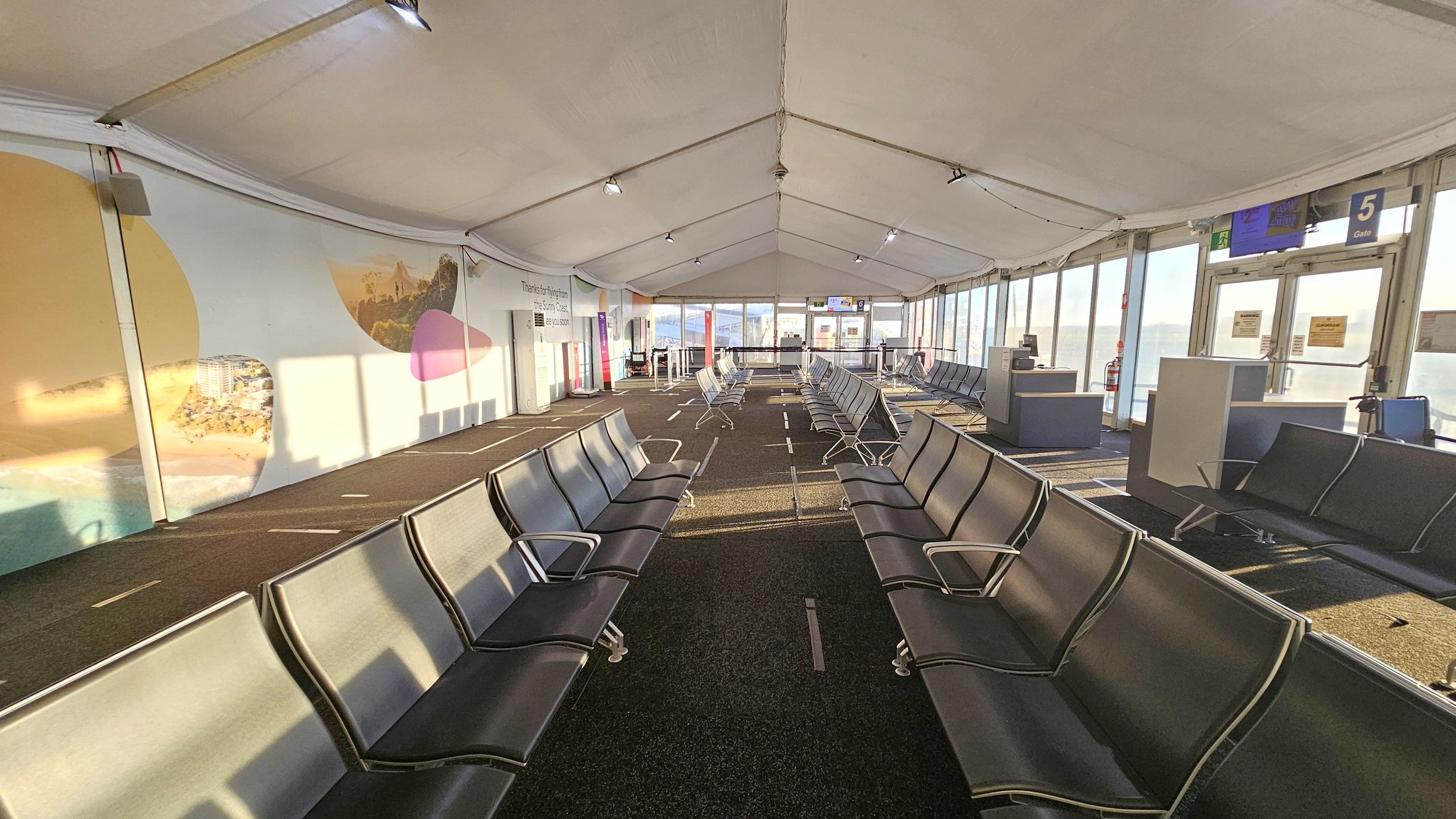 A spacious airport waiting area with rows of black seating on both sides. The left wall features large, colourful murals with abstract shapes and images of landscapes. The right side has large windows allowing natural light to flood in, with signs for Gate 5 visible. The floor is carpeted in dark tones, and the ceiling is tent-like with exposed beams and lighting fixtures. In the background, there are more seats and a boarding area with signs and barriers.