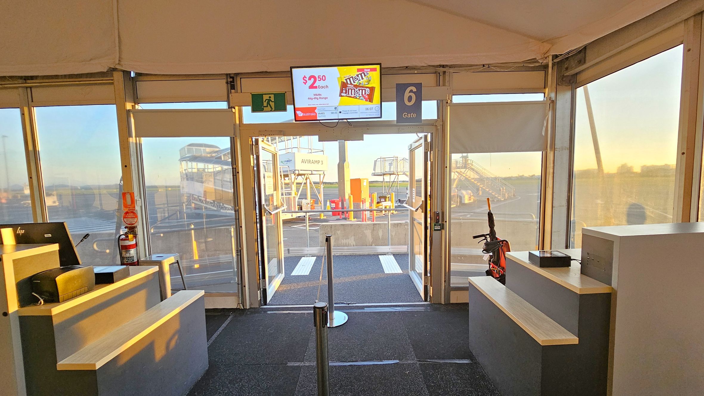 Inside an airport gate area with a carpeted floor. In the centre, glass doors lead to the tarmac, where boarding ramps are visible. Above the doors, a digital screen displays an advertisement. To the left, a counter with a computer and a fire extinguisher. On the right, another counter with a small device and an umbrella. The ceiling is tent-like, and the area is well-lit with natural light streaming through the windows.