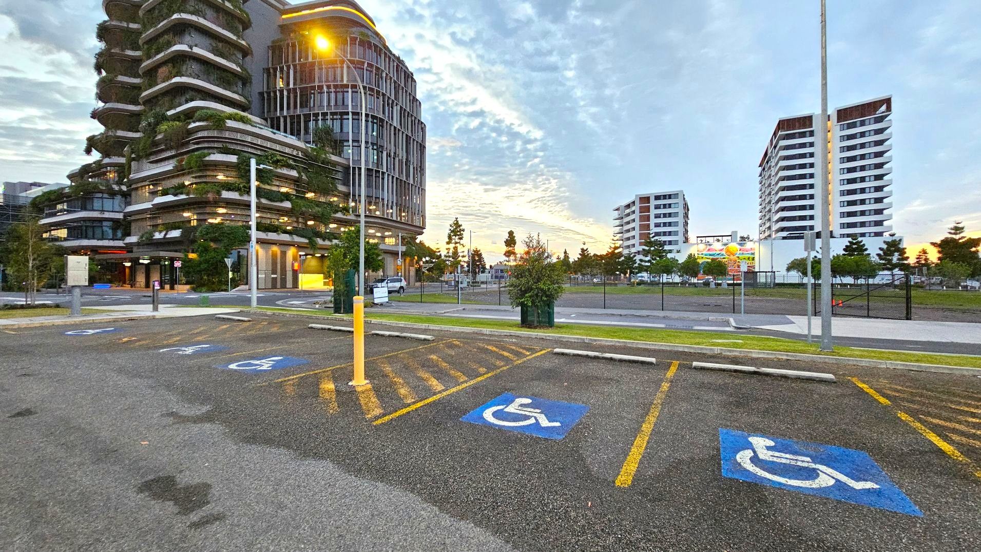 Accessible car parking. Arrival and Parking. Sunshine Coast City Hall. On the left, a modern building with curved balconies and greenery is illuminated by warm lights. In the foreground, there are marked disabled parking spaces on an asphalt surface. A tall streetlamp casts a yellow glow. In the background, two white multi-storey buildings stand against a cloudy sky. A billboard and several small trees are visible near a fenced area. The scene is urban with a mix of architecture and nature. Accessible car parking. Arrival and Parking. Sunshine Coast City Hall. On the left, a modern building with curved balconies and greenery is illuminated by warm lights. In the foreground, there are marked disabled parking spaces on an asphalt surface. A tall streetlamp casts a yellow glow. In the background, two white multi-storey buildings stand against a cloudy sky. A billboard and several small trees are visible near a fenced area. The scene is urban with a mix of architecture and nature.