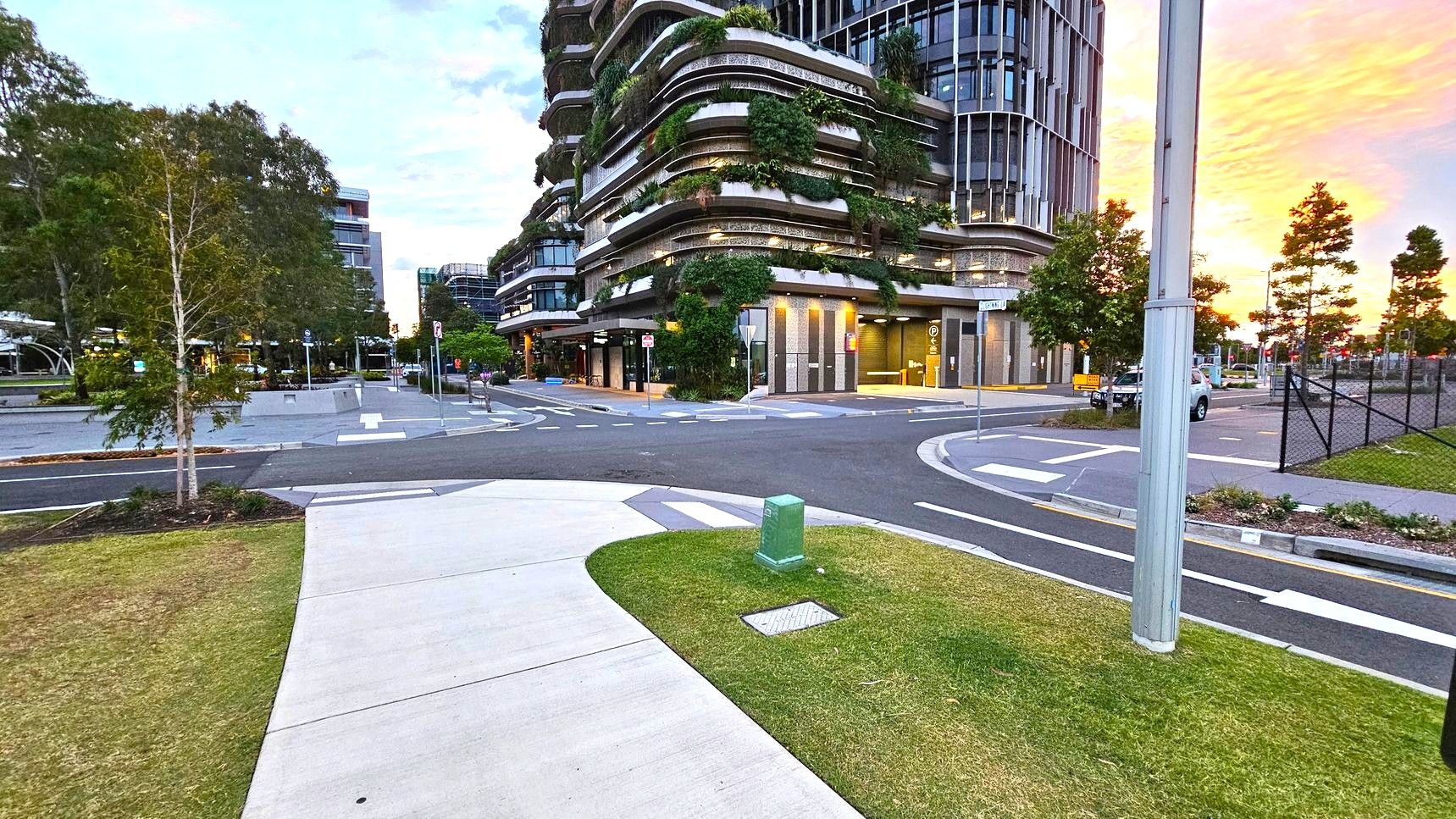 Concrete pathways. Arrival and Parking. Sunshine Coast City Hall. A modern urban scene with a curved building on the right, featuring multiple levels adorned with greenery. The foreground has a concrete path and a small patch of grass with a green utility box. On the left, there are trees and a paved area leading to more buildings. A street with marked lanes and a pole is visible, and the background shows a sunset with orange hues. Concrete pathways. Arrival and Parking. Sunshine Coast City Hall. A modern urban scene with a curved building on the right, featuring multiple levels adorned with greenery. The foreground has a concrete path and a small patch of grass with a green utility box. On the left, there are trees and a paved area leading to more buildings. A street with marked lanes and a pole is visible, and the background shows a sunset with orange hues.