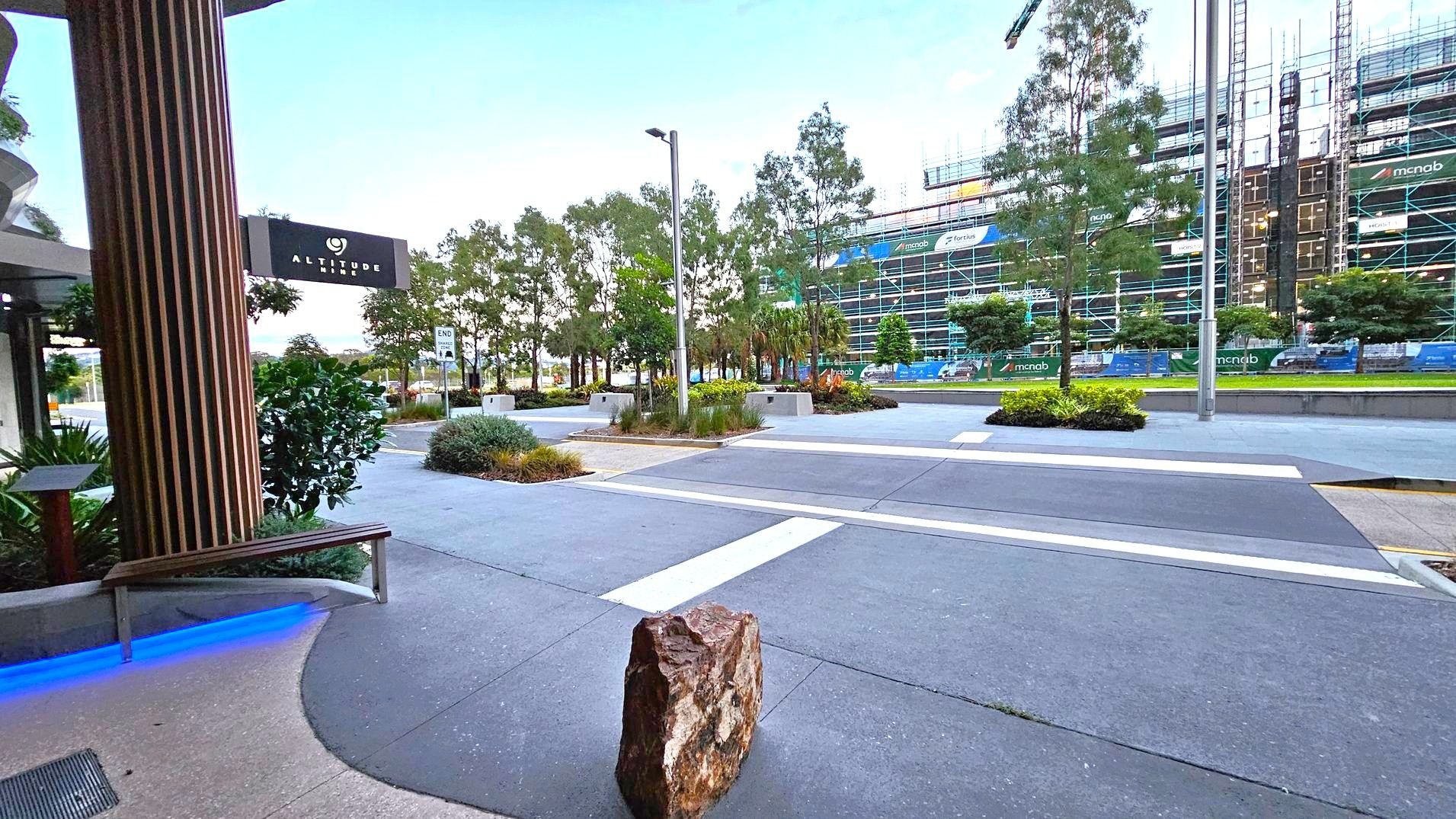 Street view. Arrival and Parking. Sunshine Coast City Hall. View from the Sunshine Coast City Hall entrance looking across First Avenue. A pedestrian crossing extends across the road directly ahead, with landscaped areas, trees and multi-storey buildings visible on the opposite side. A blue-lit bench and part of the covered entrance are visible on the left. Street view. Arrival and Parking. Sunshine Coast City Hall. View from the Sunshine Coast City Hall entrance looking across First Avenue. A pedestrian crossing extends across the road directly ahead, with landscaped areas, trees and multi-storey buildings visible on the opposite side. A blue-lit bench and part of the covered entrance are visible on the left.