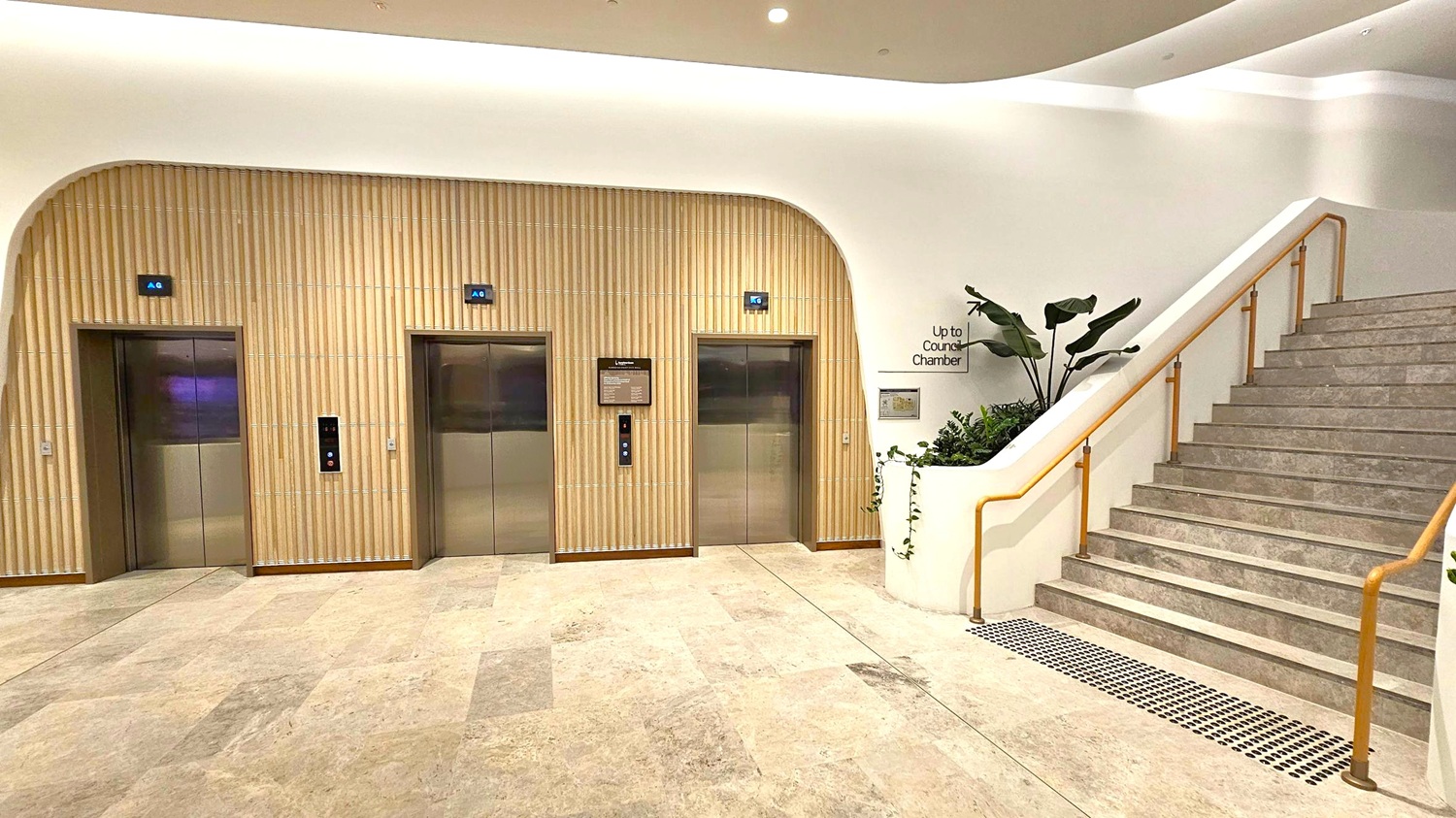 On the left, there are two lift doors with digital floor indicators above them, set in a wall with vertical wooden panelling. In the middle, a sign and lift buttons are visible between the lifts. On the right, a staircase with a wooden handrail leads upwards, accompanied by a planter with green plants. The floor is tiled in a light stone pattern, and the ceiling is smooth with recessed lighting.