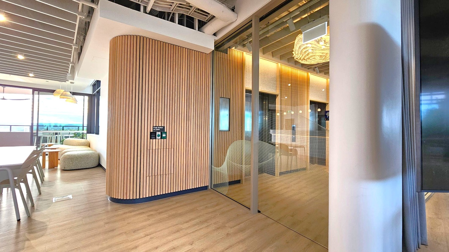 A modern interior space with a wooden slatted wall on the left, featuring a push button for door access. A glass partition with a decorative pattern separates the area. On the left, a seating area with cushioned chairs and a table is visible, along with large windows offering an outside view. The ceiling has exposed beams and hanging lights. The floor is wooden, and a large column is on the right.