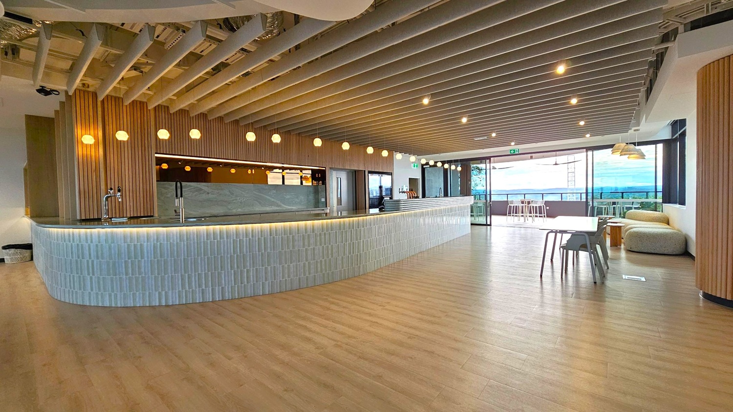 A modern interior with a long, curved bar counter on the left, featuring a tiled front and a dark countertop with a tap. Above, round pendant lights hang from a ceiling with exposed beams. On the right, a seating area includes a table with chairs and a cushioned bench. Large windows in the background offer a view outside, and the floor is wooden.