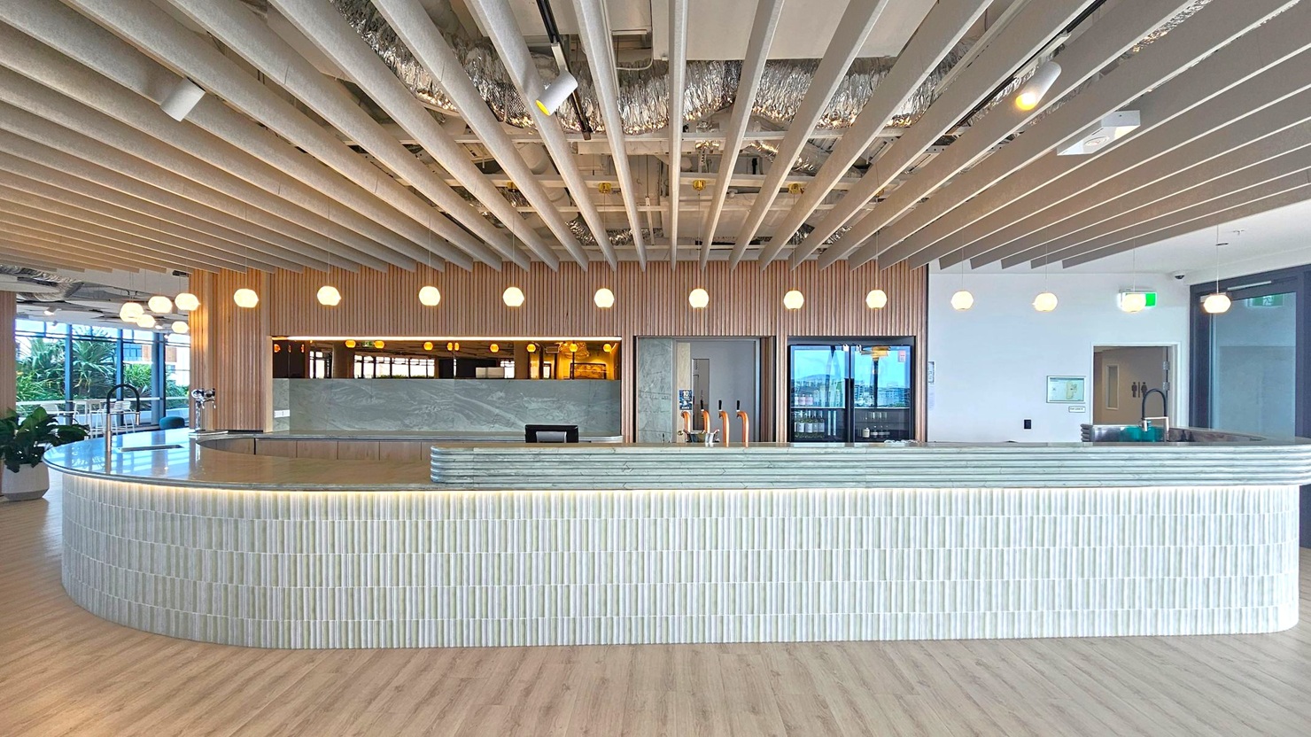A modern interior space with a large, curved bar counter in the foreground, covered in light-coloured tiles. The ceiling features exposed beams and ductwork, with round pendant lights hanging evenly across the space. In the background, there is a wooden panelled wall with a marble counter and taps. To the left, large windows allow natural light in, and there are some plants visible. The floor is wooden, complementing the overall neutral and natural colour scheme.