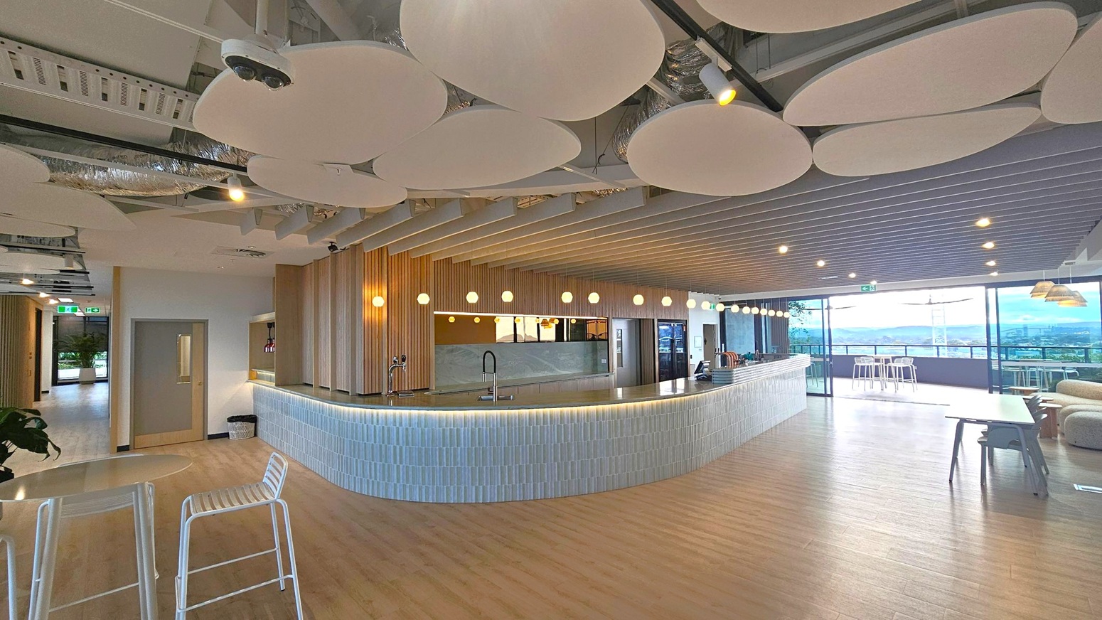 A modern interior space with a curved bar counter in the centre, featuring a tiled front and a sleek countertop with taps. Above, round light fixtures hang from a ceiling adorned with circular acoustic panels and exposed ductwork. On the right, large glass windows offer a view of the outdoors, with high stools and tables nearby. The left side has a corridor leading to another area. The floor is wooden, and the space is open and airy.