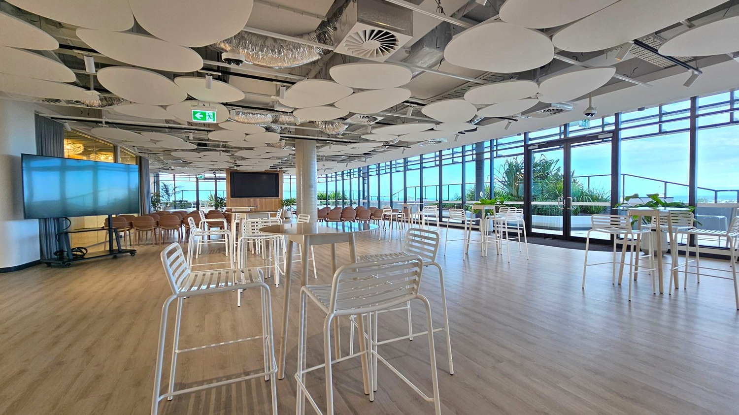 A spacious room with light wooden flooring and a ceiling adorned with circular acoustic panels. On the left, there's a large screen on a stand and several wooden chairs. In the centre, white metal high tables and chairs are scattered throughout. The right side features large windows and glass doors, offering a view of greenery outside. The room is well-lit with natural light streaming in.