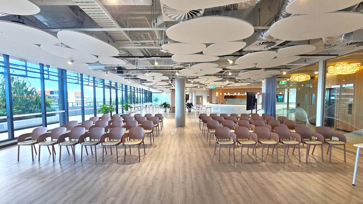 A modern conference room with rows of brown chairs arranged in the centre on a light wooden floor. The left side features large glass windows with views of greenery and buildings outside. The right side has a bar area with high stools and a wooden wall. The ceiling is adorned with circular acoustic panels and exposed ductwork. Potted plants are placed throughout the space, adding a touch of greenery indoors.