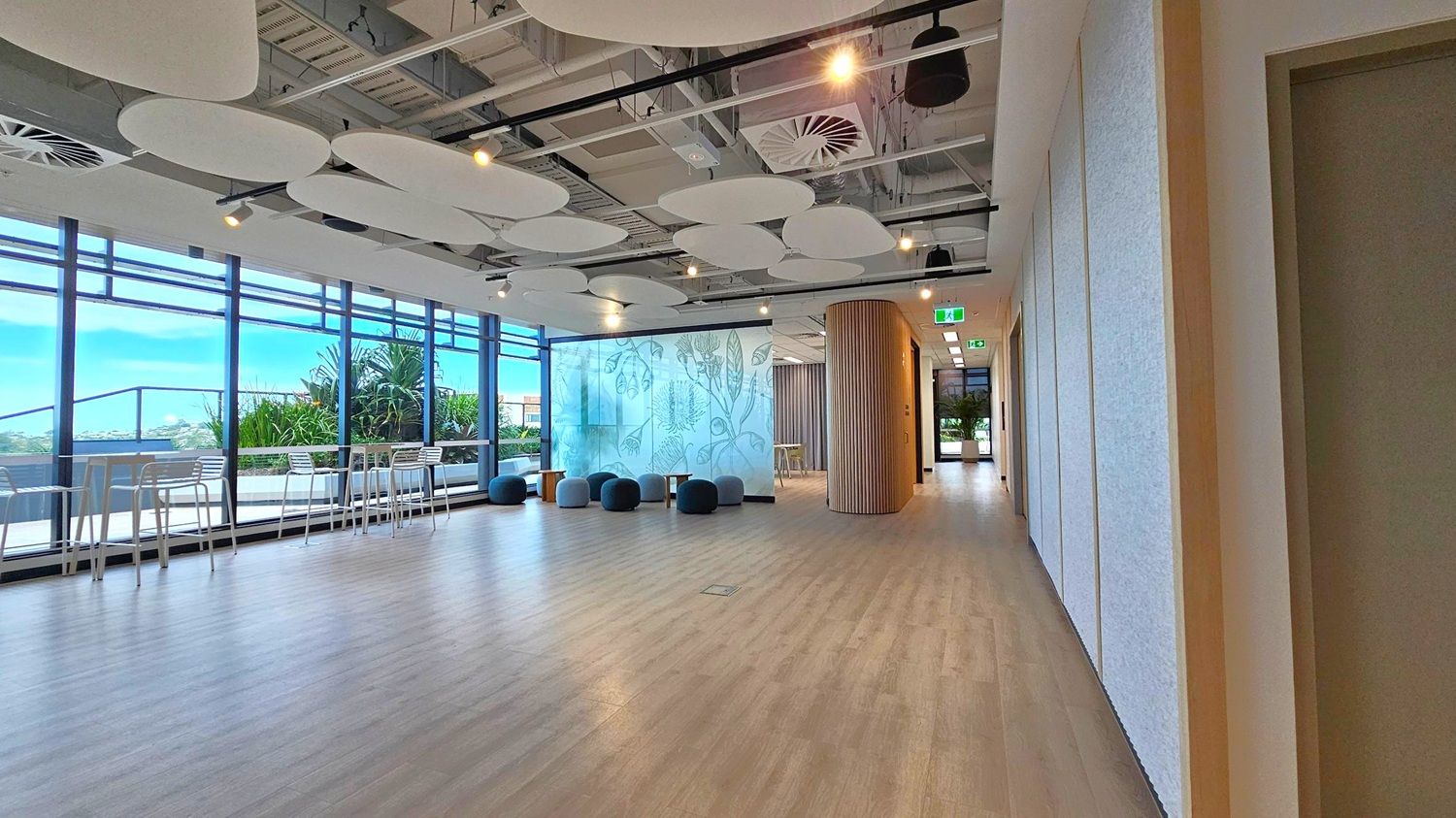 A spacious, modern interior with light wooden flooring. On the left, large windows offer a view of greenery and outdoor seating with high tables and chairs. In the centre, a decorative glass wall with botanical designs is flanked by round, cushioned stools. The ceiling features circular acoustic panels and exposed ductwork. On the right, a hallway leads to another area, with a wooden column and exit signs visible.