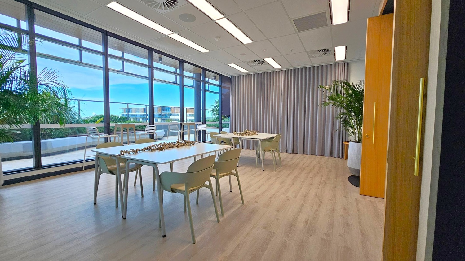 A room with large floor-to-ceiling windows on the left, offering a view of a terrace with plants and outdoor seating. Inside, there are several tables with chairs, and decorative items on the tables. The right side features a grey curtain and a large potted plant. The floor is light wood, and the ceiling has recessed lighting and ventilation panels.