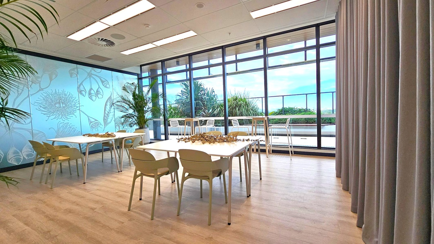 A room with light wooden flooring and a ceiling with recessed lighting. On the left, there are tables with beige chairs and a decorative arrangement on top. The wall features a botanical design. In the middle, large windows offer a view of an outdoor area with plants and white chairs. On the right, there are grey curtains.
