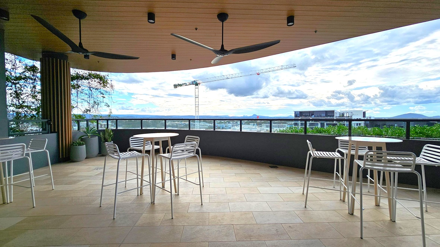 A rooftop terrace with a tiled floor and a wooden ceiling featuring two large ceiling fans. On the left, there are potted plants and a round table with white chairs. In the centre, another round table with chairs is positioned near a railing. The background shows a cityscape with a construction crane and distant mountains. The terrace is partially enclosed with a glass barrier, providing a view of the surroundings.