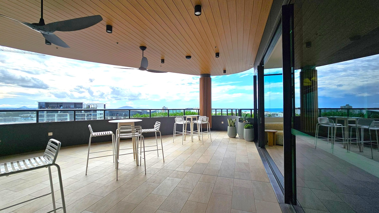 A spacious outdoor terrace with a wooden ceiling featuring ceiling fans and recessed lights. The floor is tiled in a light grey pattern. On the left, there are metal chairs and tables, while the right side has a glass wall reflecting the terrace. In the background, a railing overlooks a cityscape with buildings and greenery. Large potted plants are placed near the glass wall.