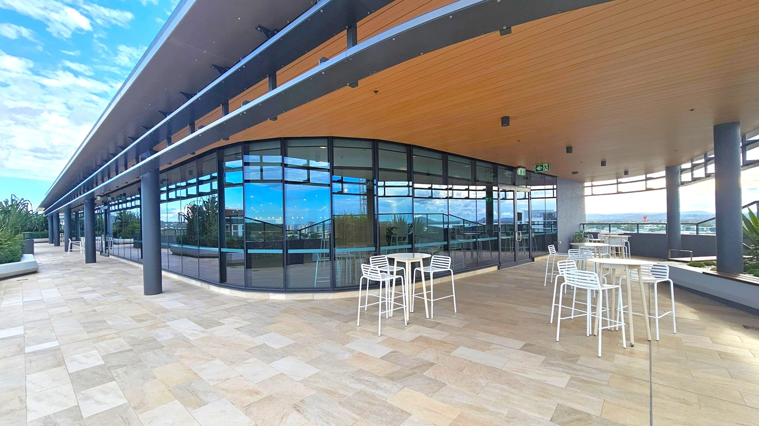 A modern outdoor terrace with a light-coloured tiled floor and a wooden ceiling. On the right, there are several white tables and chairs. The left side features a building with large glass windows reflecting the surroundings. Black columns support the overhanging roof. In the background, there are plants and a view of a distant cityscape.