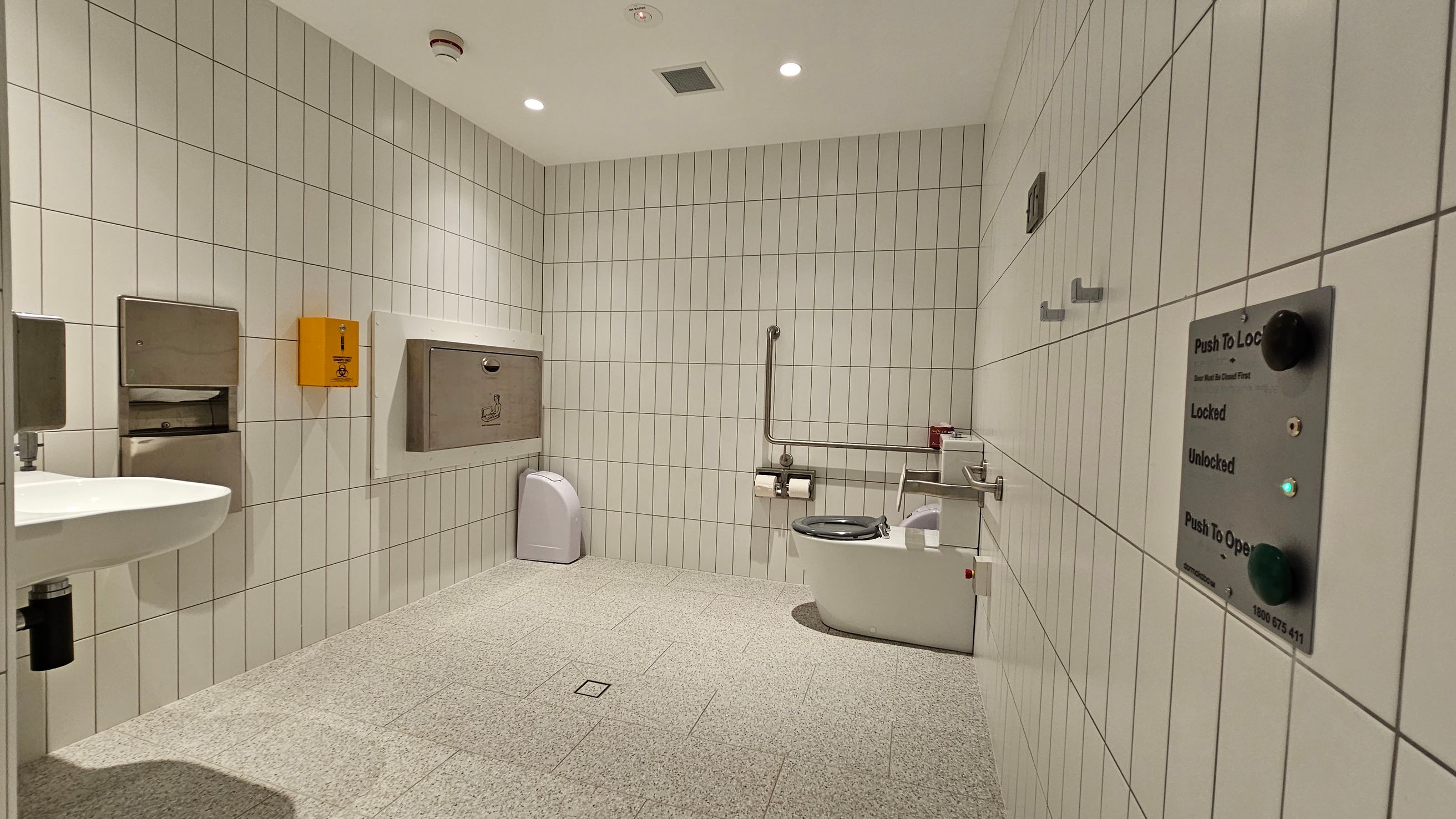 A spacious public restroom with white tiled walls and a grey tiled floor. On the left, a white sink is mounted on the wall with a stainless steel paper towel dispenser and a yellow emergency button nearby. In the centre, a baby changing station is attached to the wall above a small bin. On the right, a toilet with a support rail and a shelf holding toilet paper and a small item. A control panel for locking and unlocking the door is visible on the right wall. The ceiling has recessed lighting.