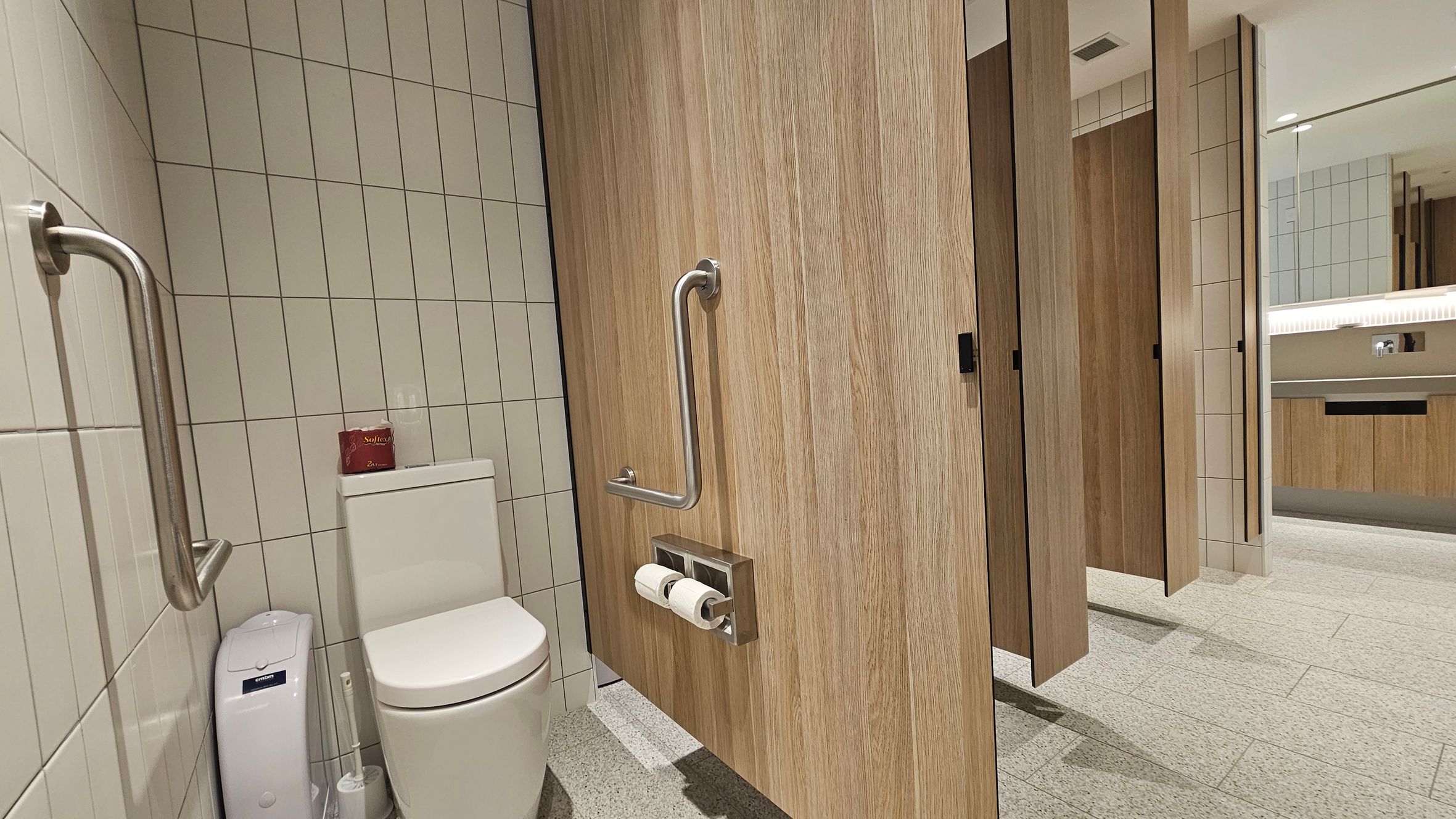 A public restroom with a tiled floor and walls. On the left, a toilet with a white tank and a small red box on top. A metal grab bar is mounted on the wall beside the toilet. A wooden door with a metal handle and toilet paper holder is partially open. On the right, a row of similar wooden doors leads to other stalls. In the background, a large mirror reflects the restroom's interior, and a countertop with a sink is visible.
