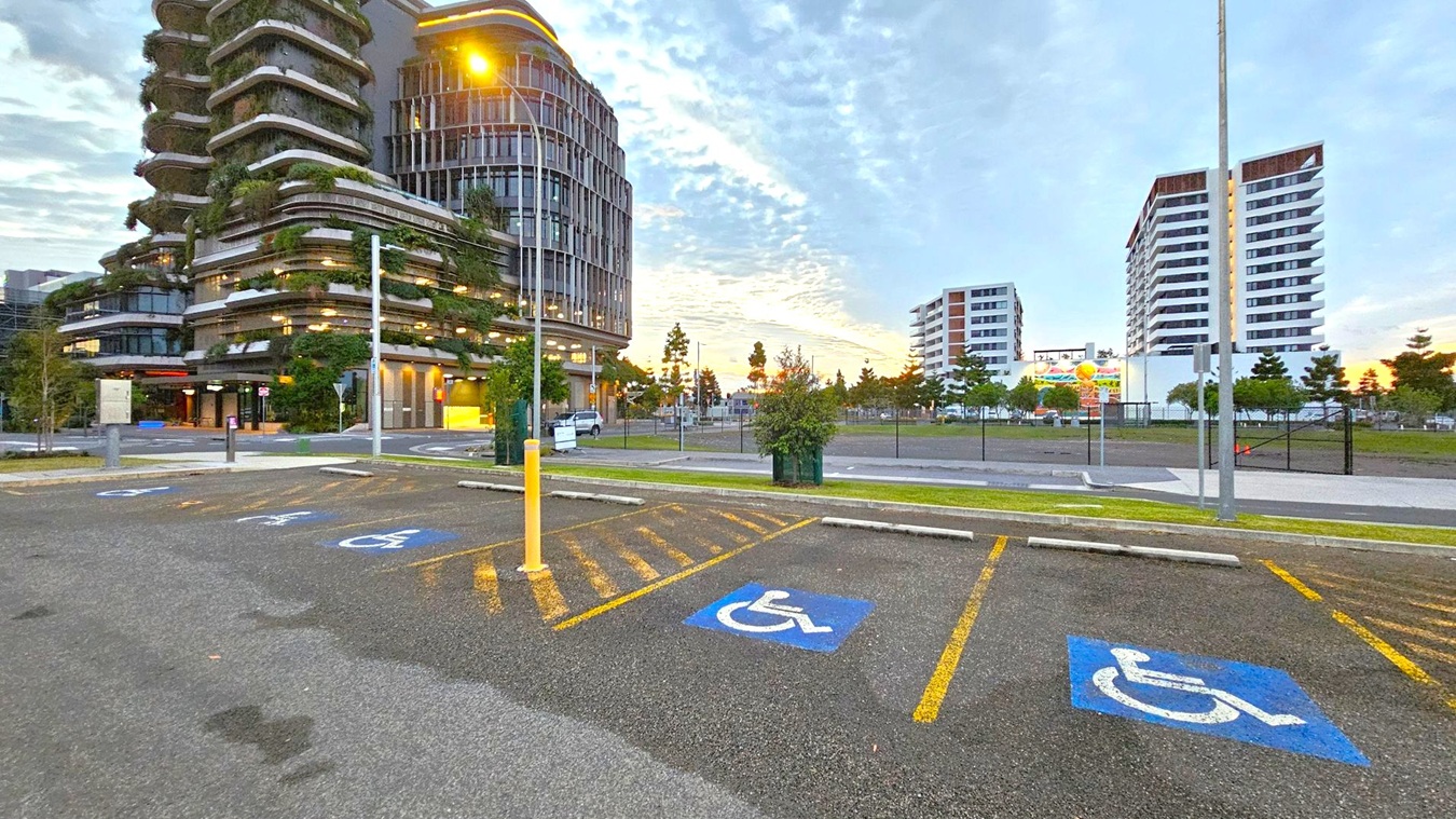 In the foreground, there are parking spaces marked with blue disabled signs. A yellow bollard stands near the centre. On the left, a modern building with curved balconies and greenery is visible. In the background, two white multi-storey buildings are on the right. Trees and a billboard are situated between the buildings. The ground is asphalt, and a streetlamp is lit, casting a warm glow.
