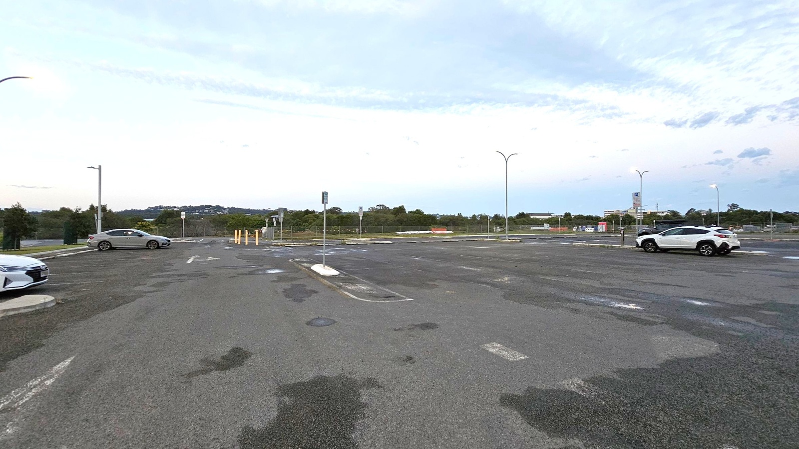 An empty car park with a few vehicles scattered around. On the left, a silver car is parked near a streetlight. On the right, a white SUV is parked under another streetlight. The ground is asphalt with visible patches and markings. In the background, there are trees and some distant buildings. Several streetlights are lit, casting a soft glow over the area. The sky is overcast with a hint of light.